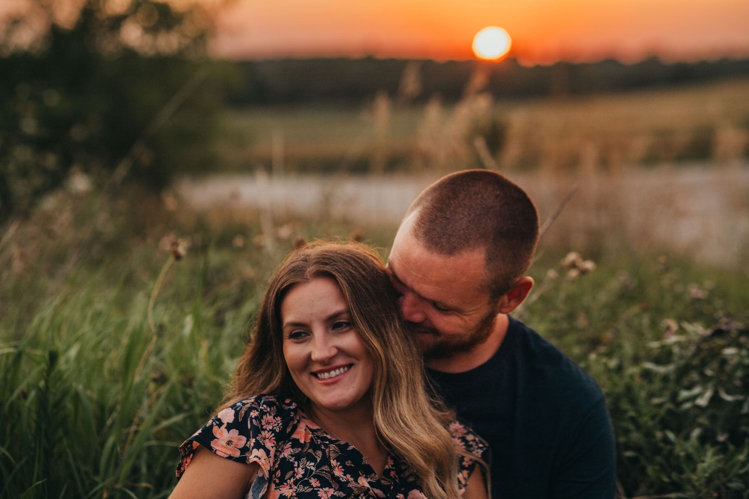 A couple shares an intimate moment in a field at sunset as the orange sun glows in the background.