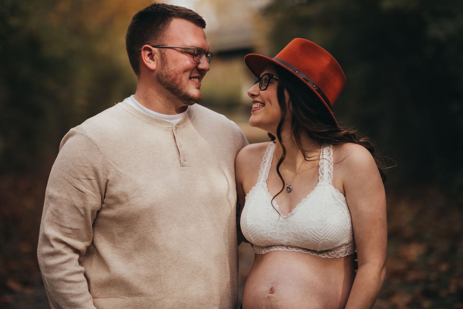 A couple smiles at each other outdoors during a maternity photoshoot in autumn, wearing casual earth-toned clothing.