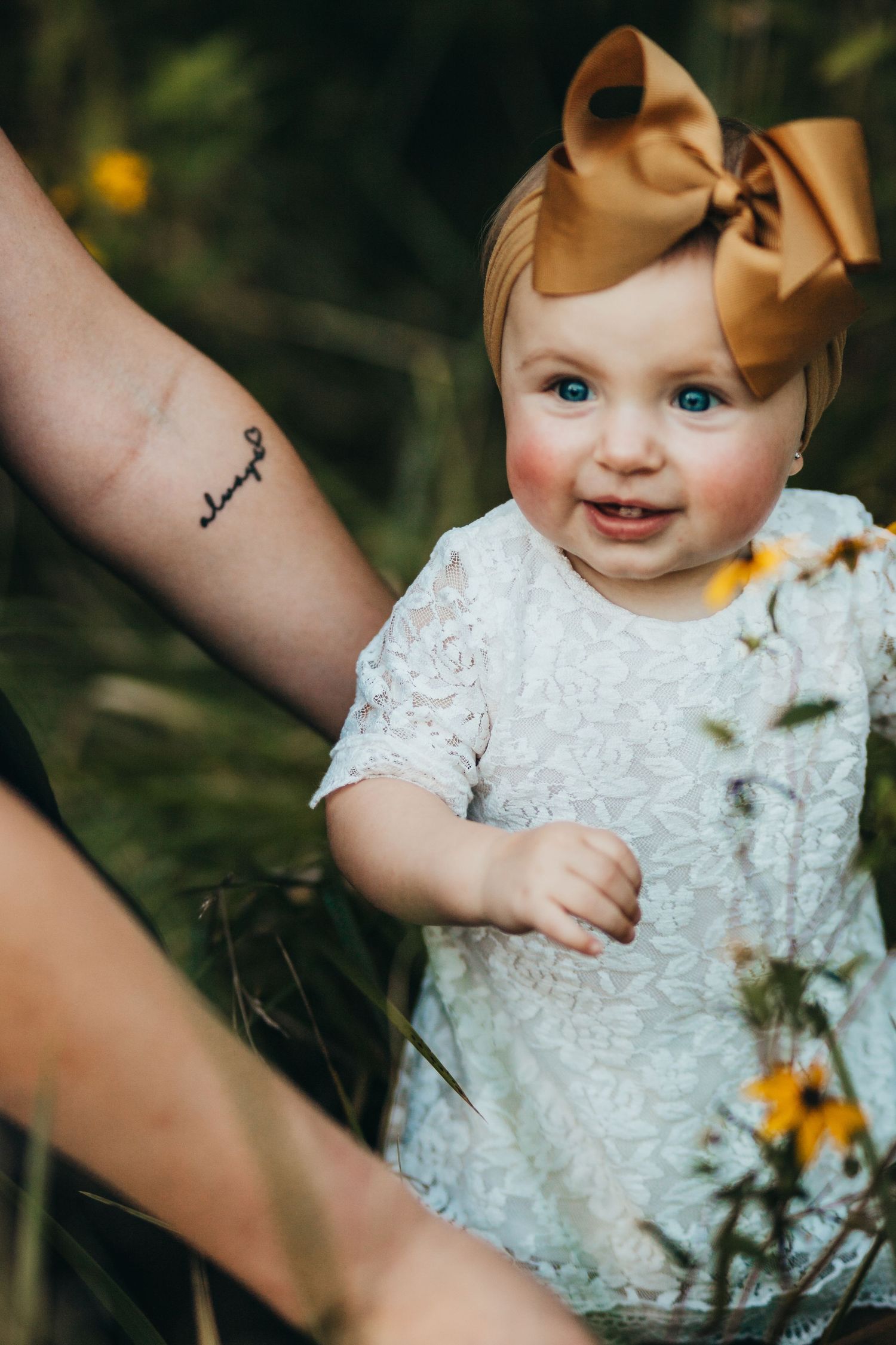 A baby wearing a white lace dress and large tan bow headband sits outdoors among wildflowers.