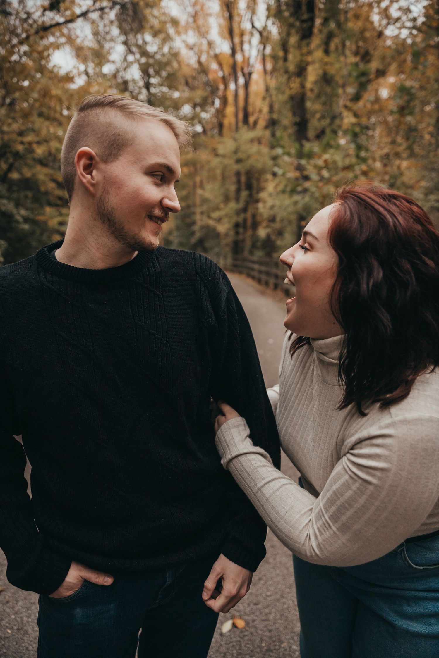 A couple in warm sweaters share a joyful moment on a fall path surrounded by autumn trees.