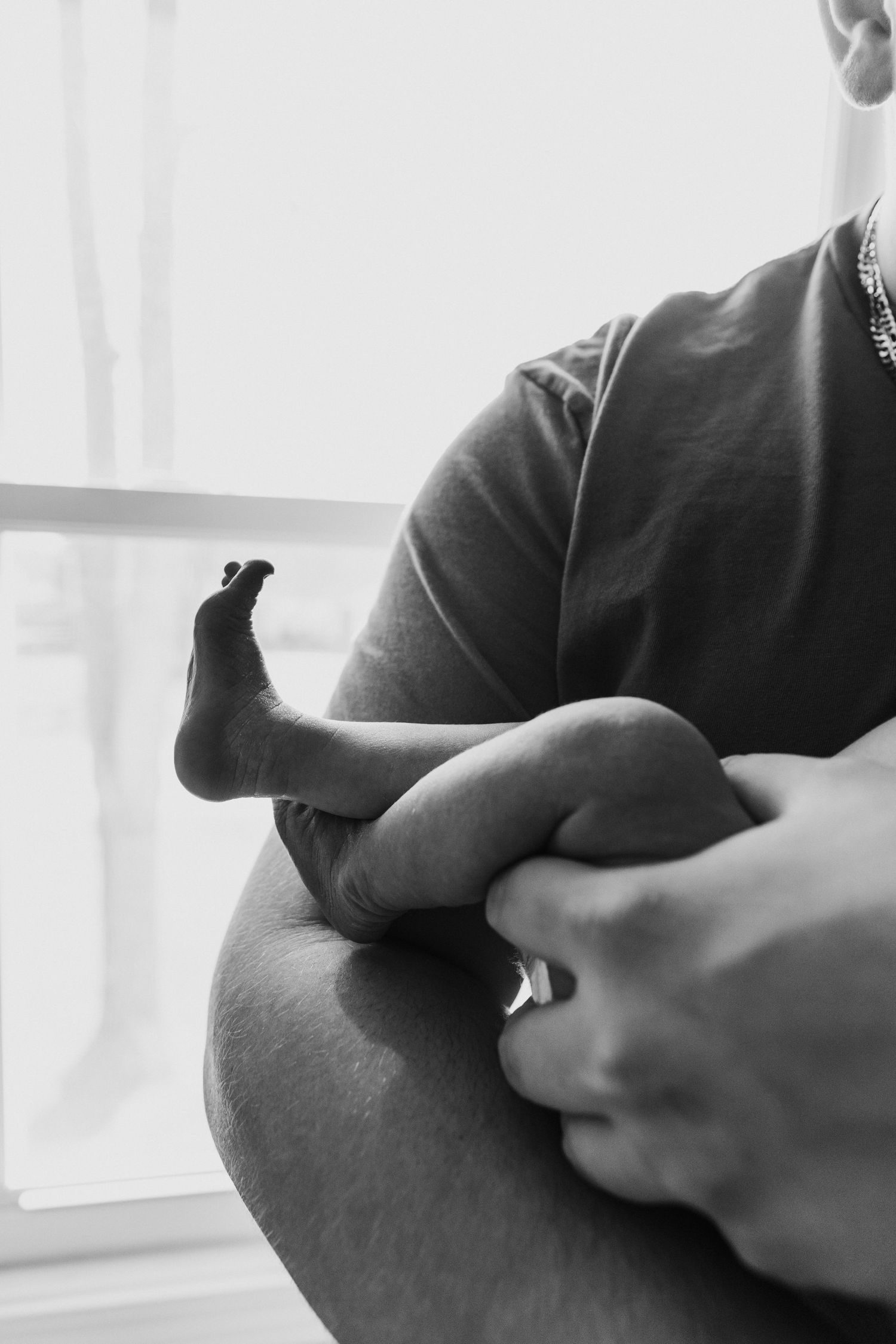 A black and white close-up photograph of an infant's tiny foot being gently held by adult hands near a window.