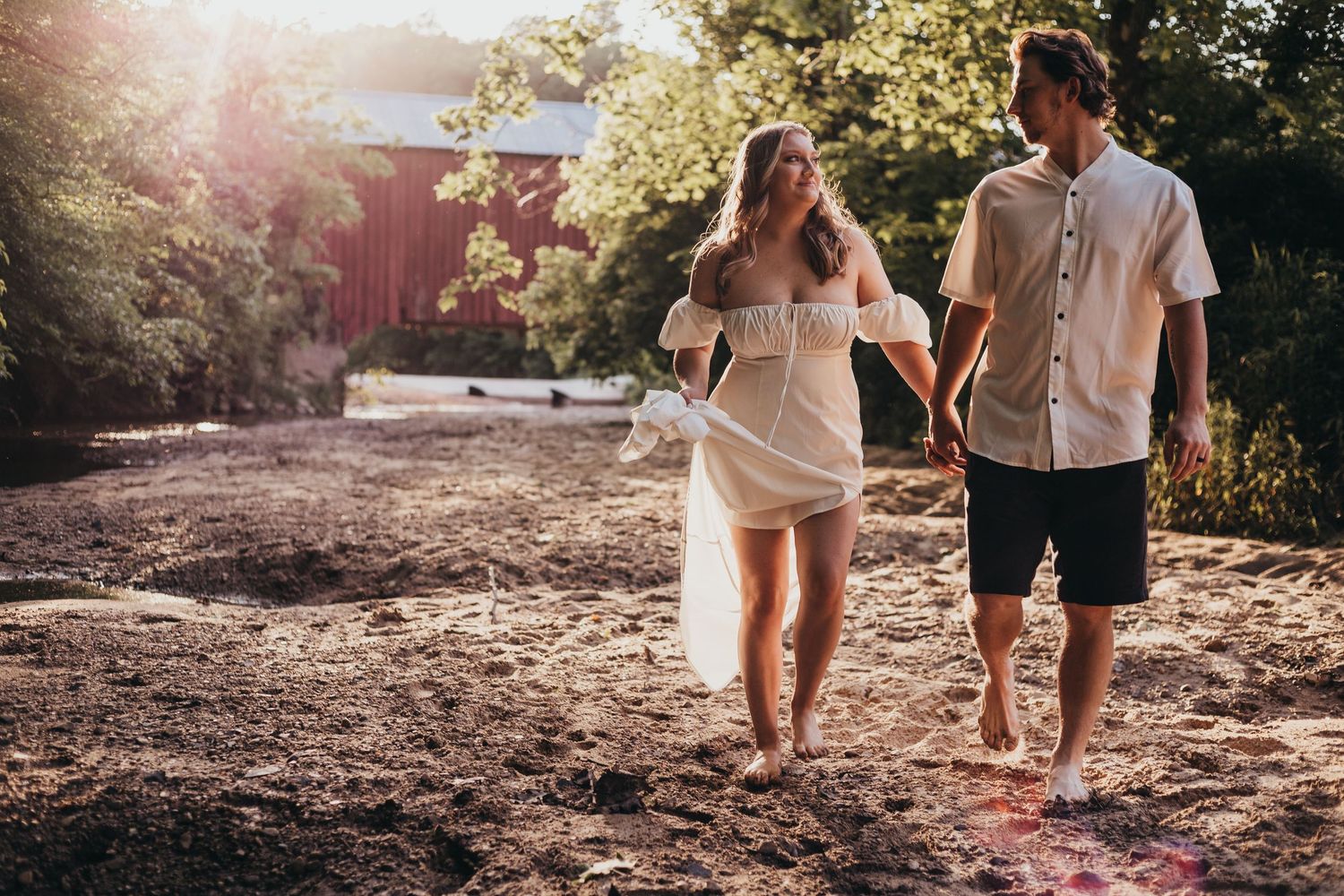 Couple walking barefoot on dirt path in summer sunlight with sunflare and trees in background.