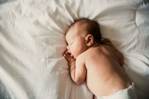 Sleeping newborn baby lying on white bedding with bare back visible in natural light. Newborn photos Terre Haute