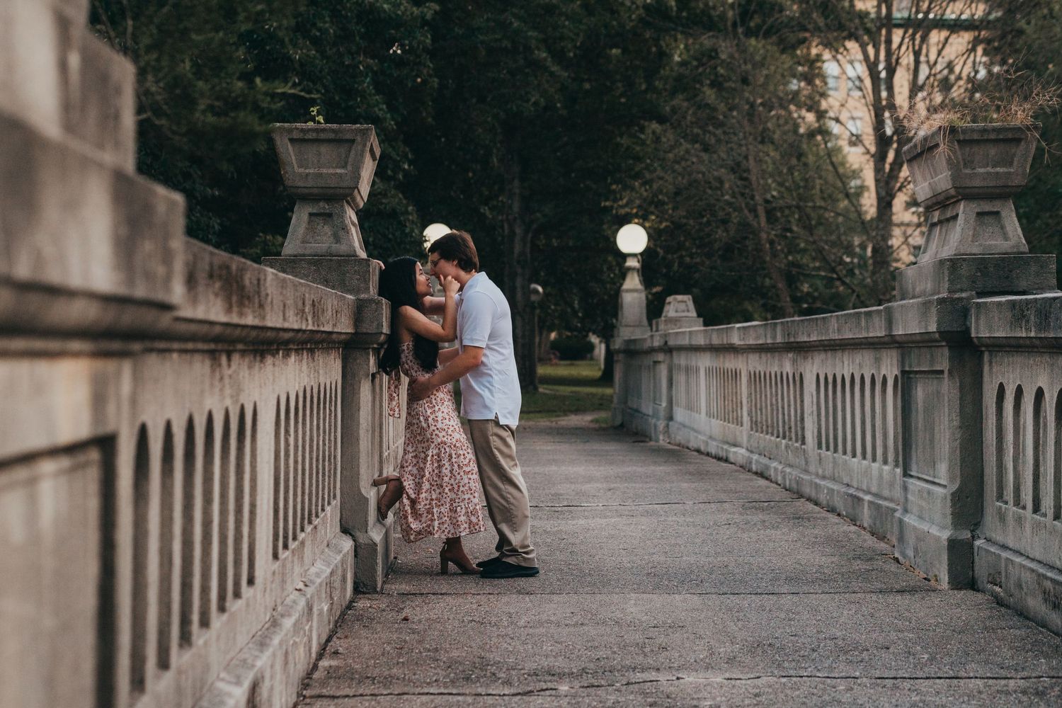 Two people share an intimate moment while embracing on a stone bridge walkway surrounded by ornate railings and lamp posts.