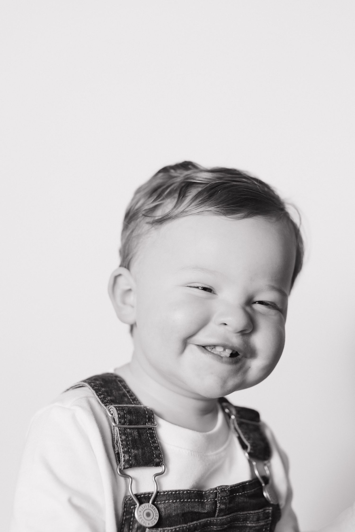 Black and white portrait of a happy toddler in denim overalls, smiling with a playful squinting expression.