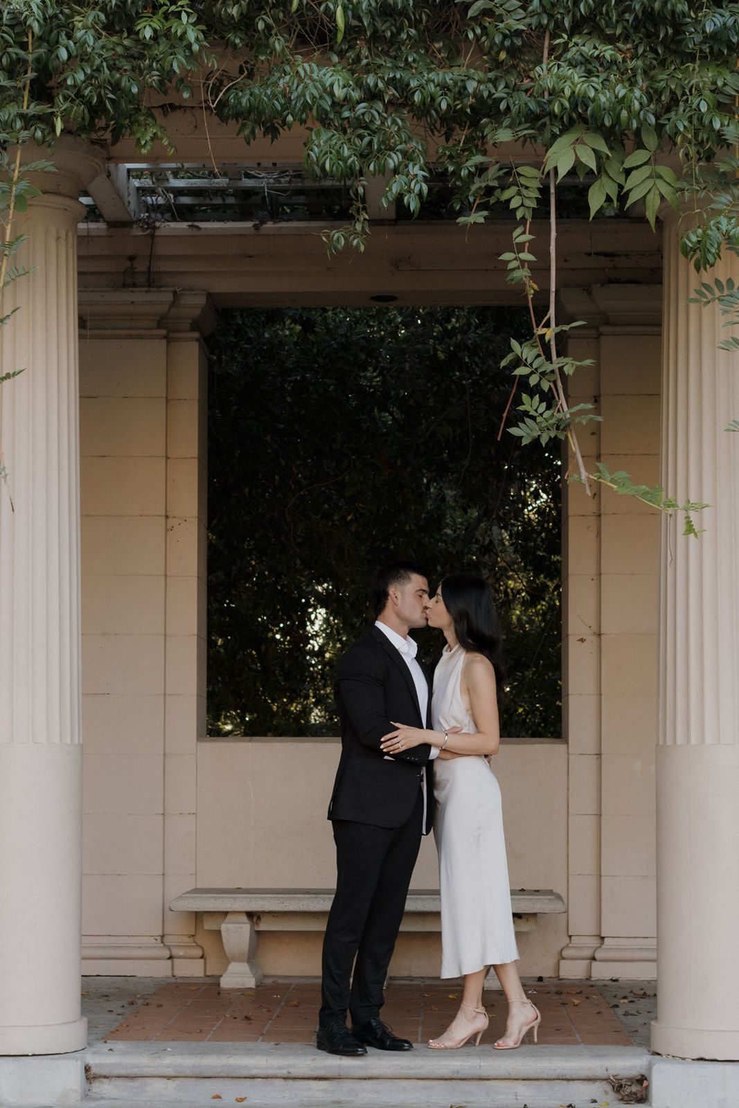 A couple shares an intimate moment in an elegant outdoor alcove with white columns and leafy vines overhead.