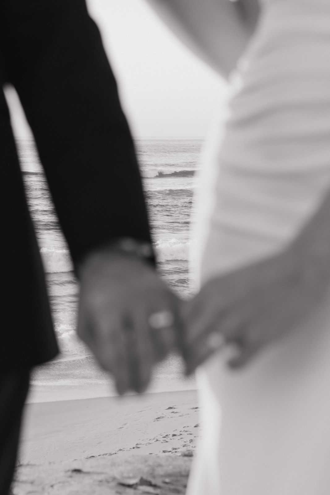 Black and white close-up of hands holding on a beach with ocean waves visible in the background.