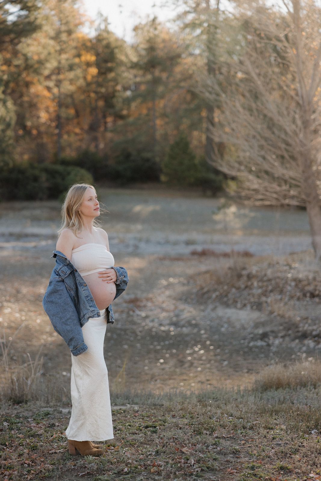 A pregnant woman in a white dress and denim jacket stands in a natural outdoor setting during autumn.
