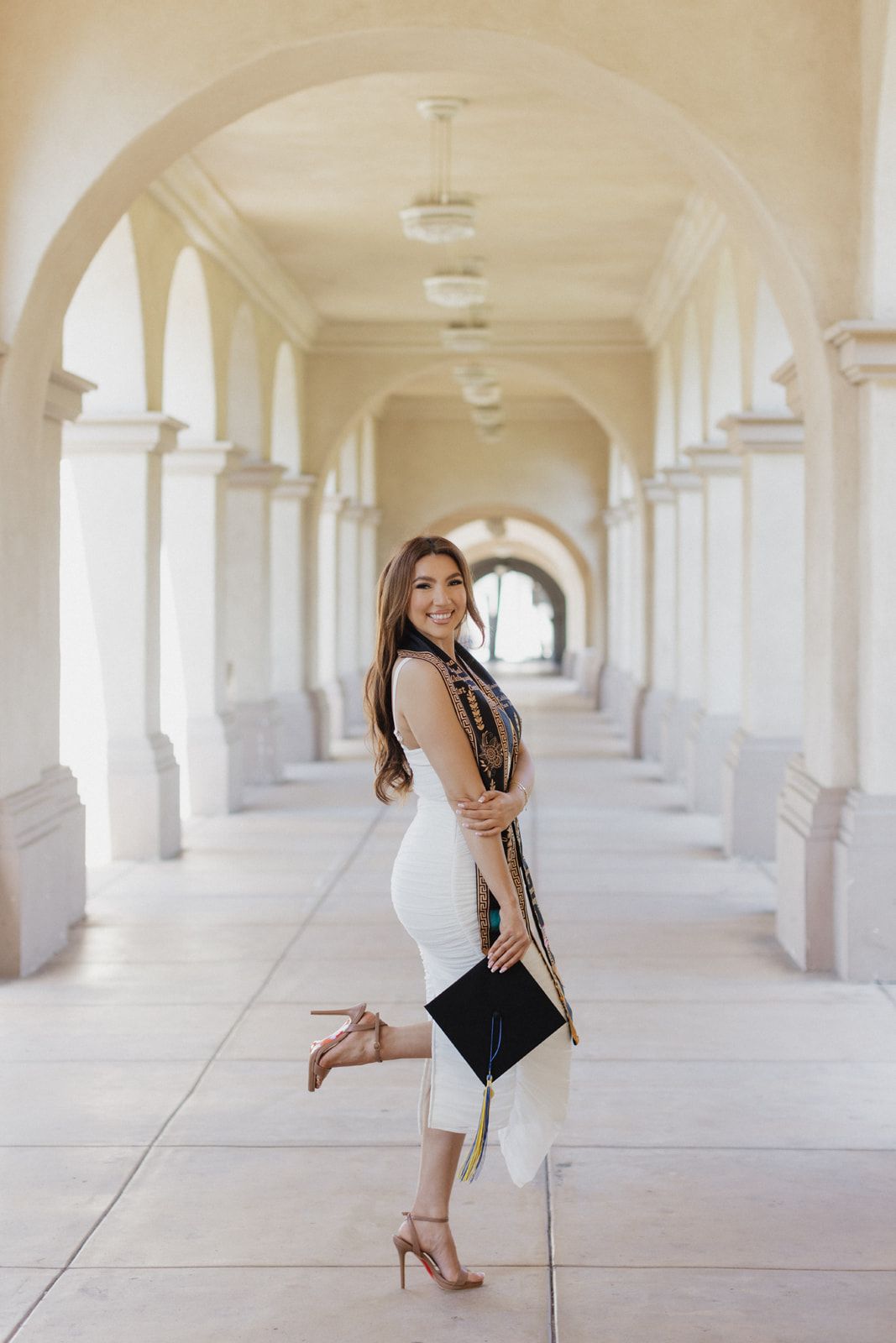 A person in a black dress poses elegantly in a bright white arched hallway with repeating columns.