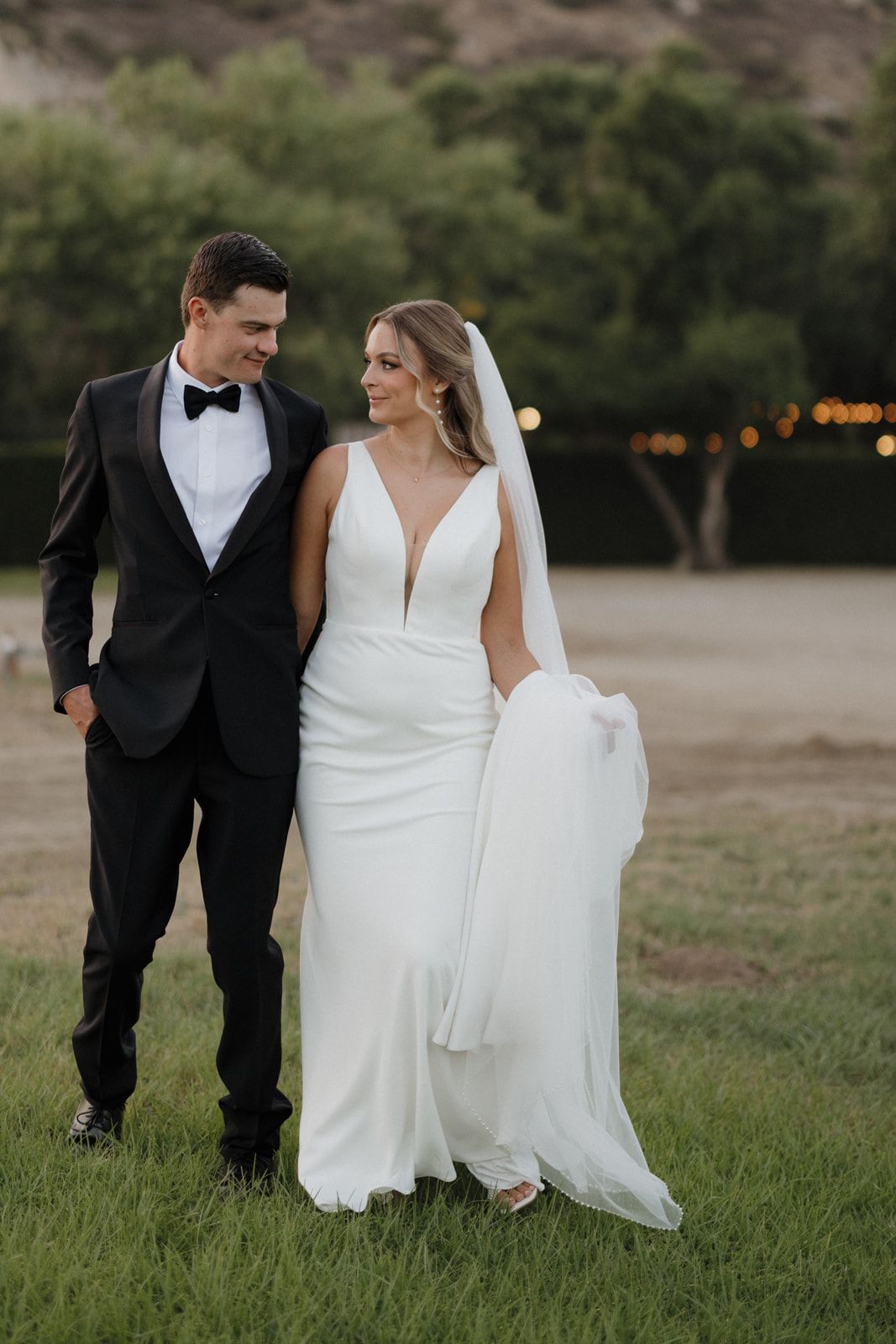 A romantic wedding couple walking together on a grassy field at dusk with string lights visible in the background.