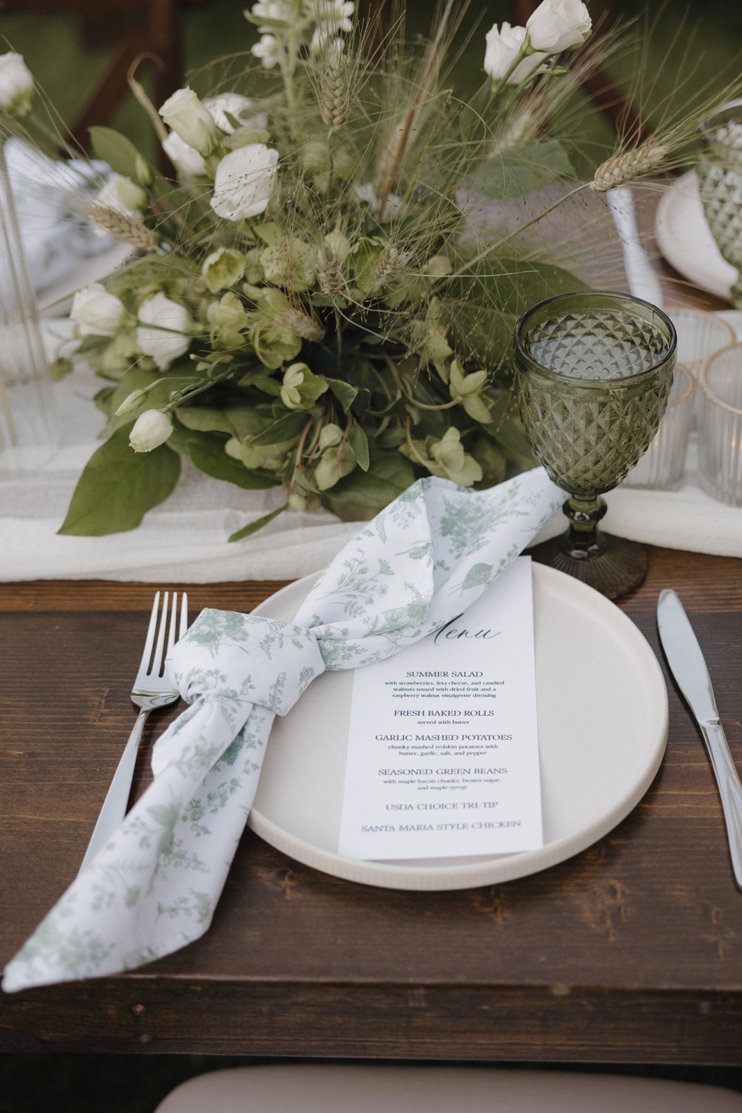 Elegant wedding place setting with white floral centerpiece, menu card, green goblet and floral napkin on wooden table.