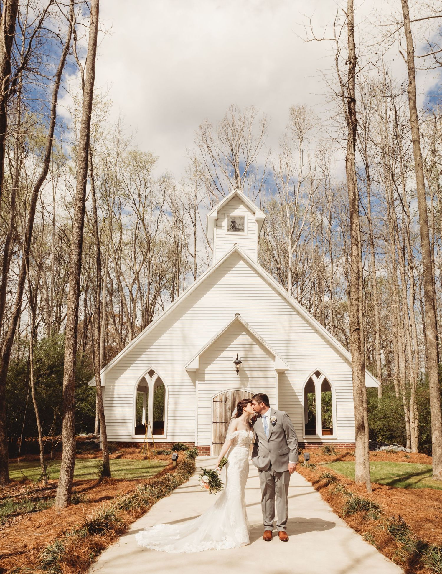 Bride and groom kissing in front of a white chapel surrounded by trees on their wedding day.