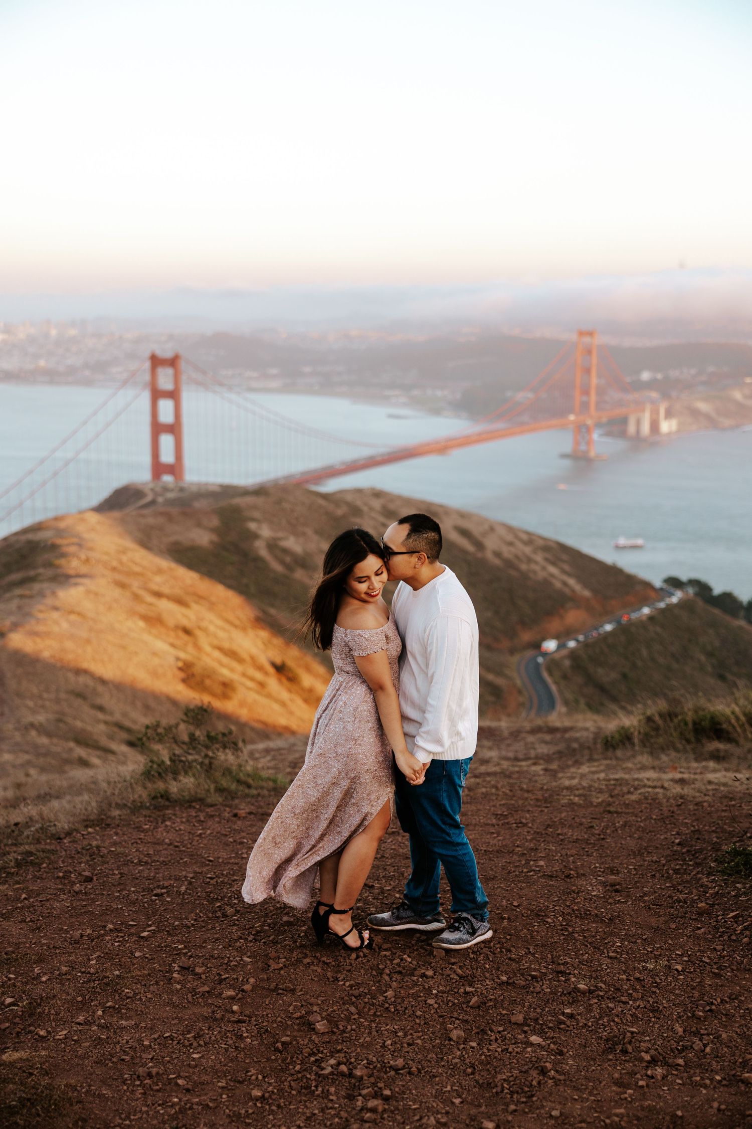couple on a mountain top with the golden gate bridge and san francisco behind them during their sunset engagement photoshoot