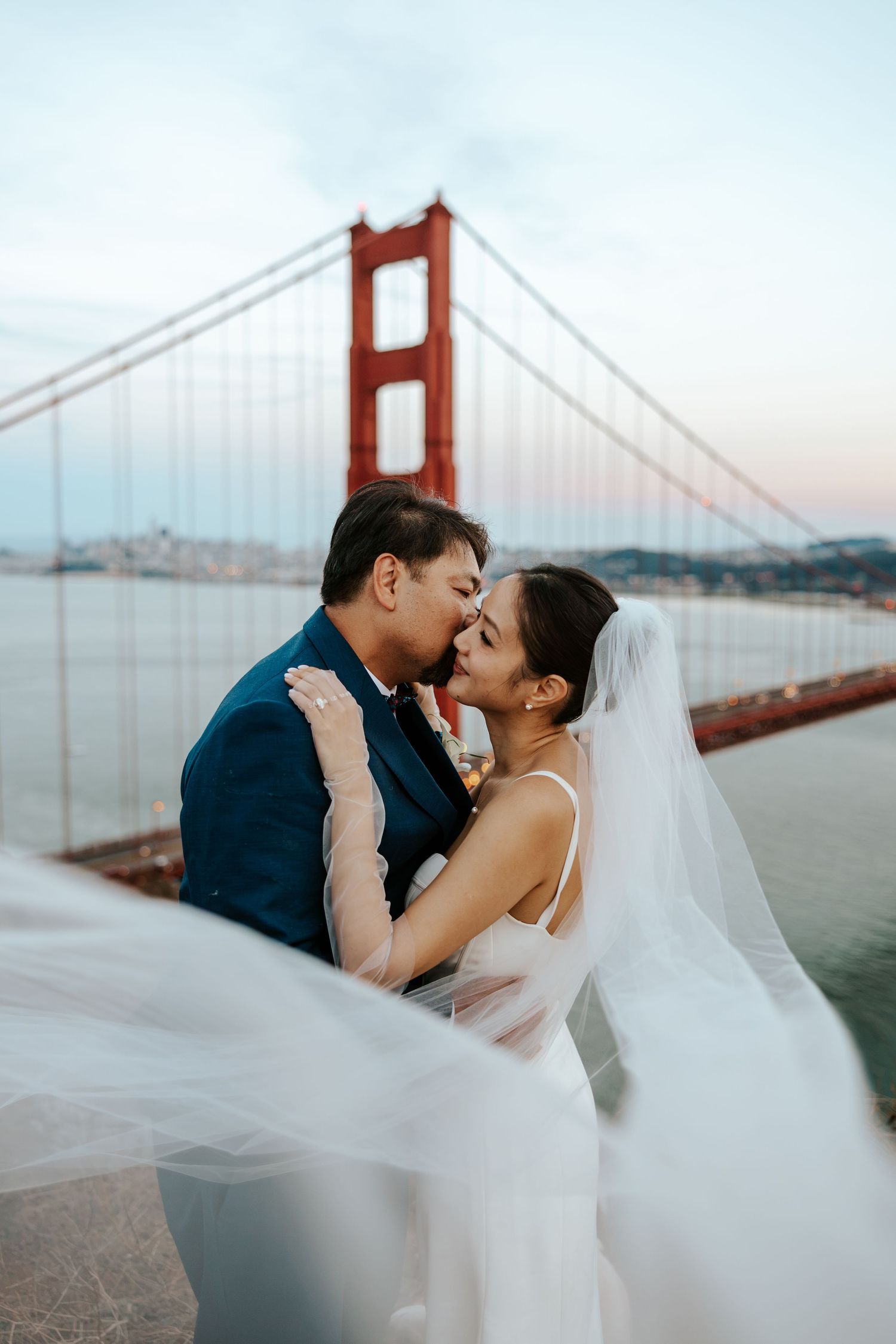 groom kissing bride's cheek while her veil flows in the wind in front of the golden gate bridge on their san francisco elopement day