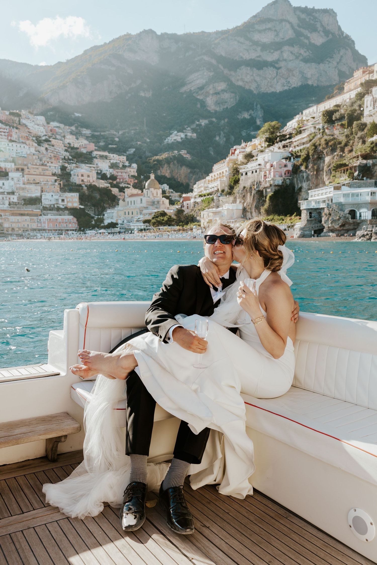 bride sitting on groom's lap kissing his cheek while sitting on a boat with a view of Positano behind them during italy elopement day