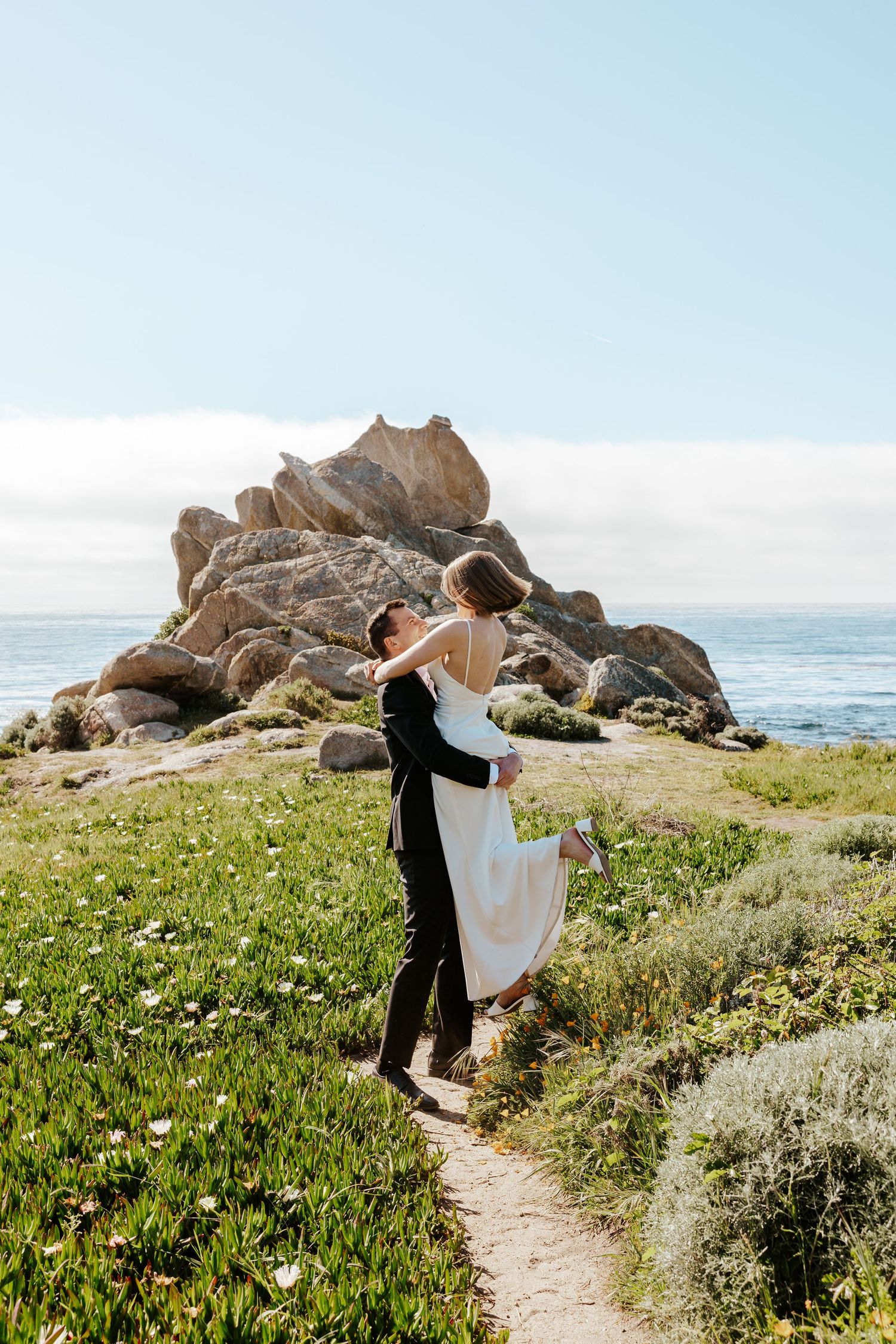 groom picking up and spinning with his bride on pathway lined by wildflowers and giant rock formation and the ocean behind them in Carmel CA
