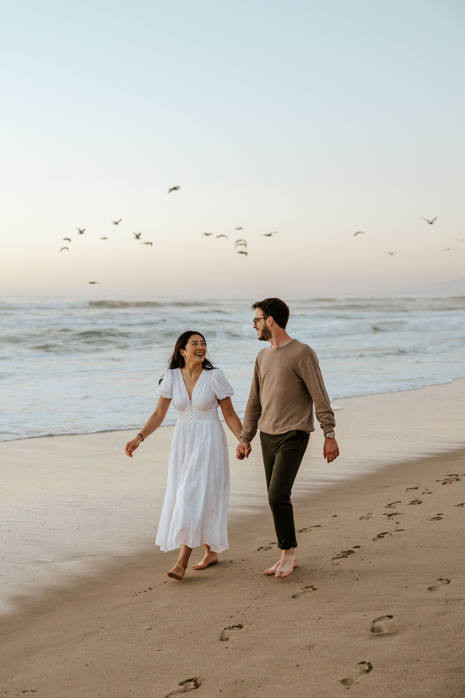 A couple walking on the beach, hand in hand, while birds fly by. A San Gregorio State Beach engagement photoshoot.