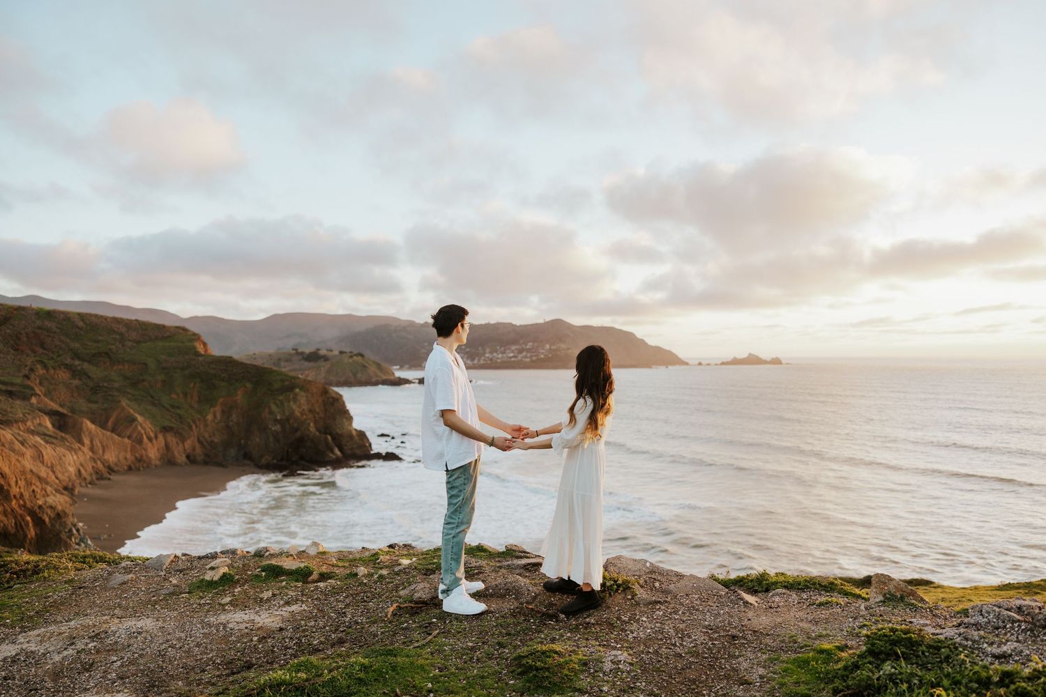 couple holding hands on a cliff top in pacifica looking out towards the ocean at sunset during their engagement photoshoot