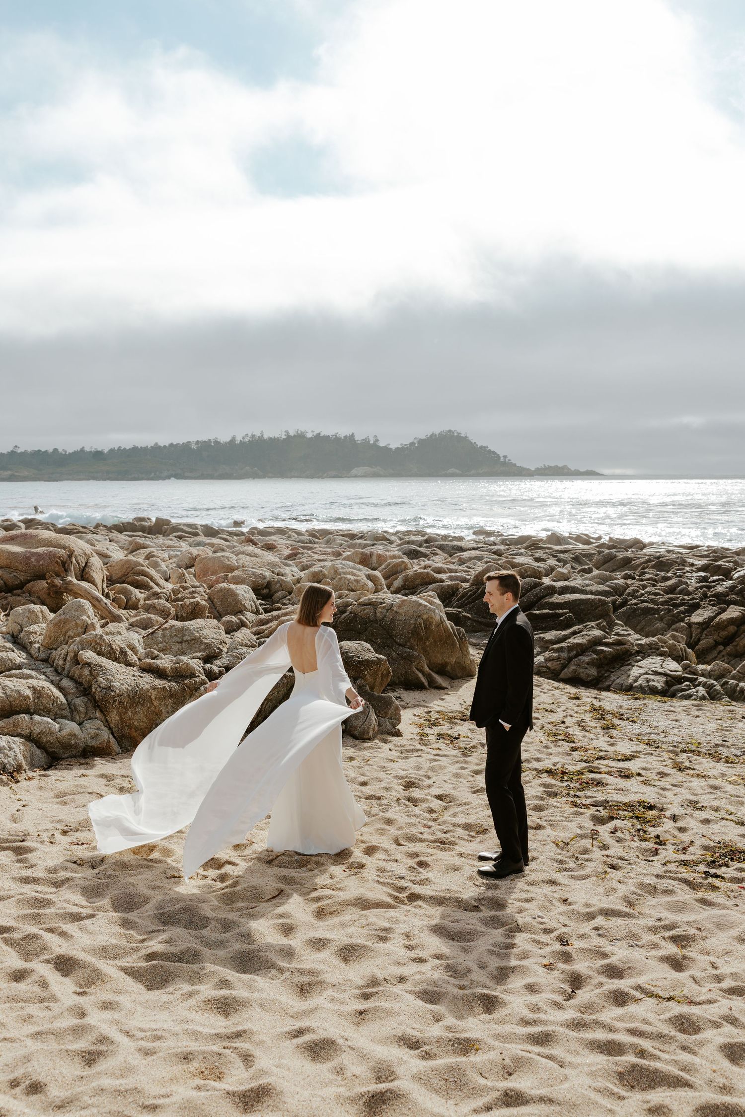 bride showing off her bridal cape to groom while on the beach during big sur elopement