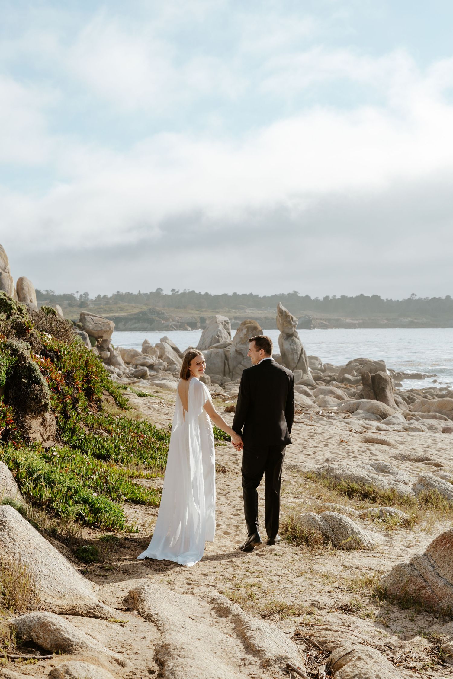 couple walking away from camera on the beach while bride looks over her shoulder