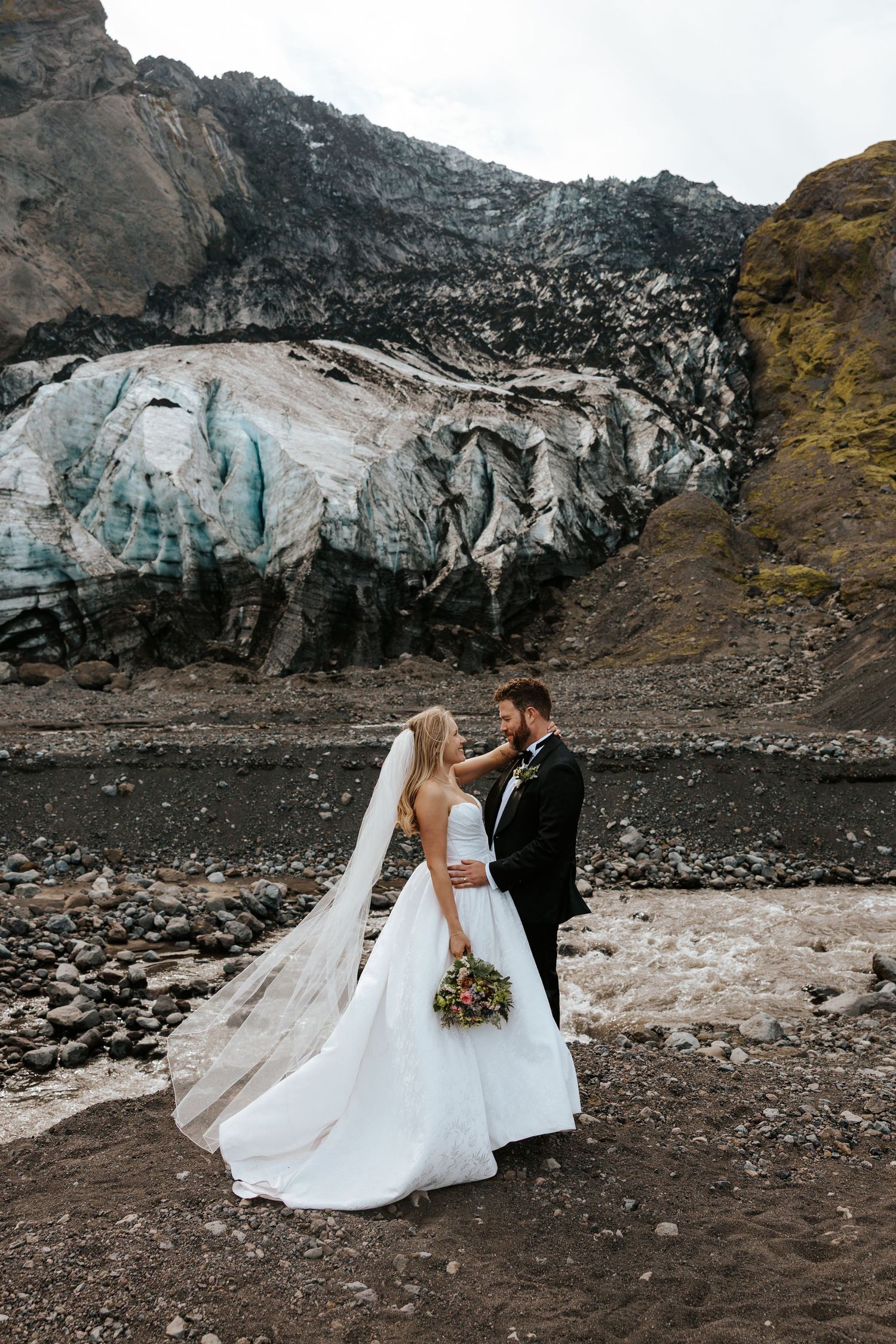 romantic photo of bride and groom staring into each other's eyes with a glacier in a mountain behind them during Iceland adventure elopement