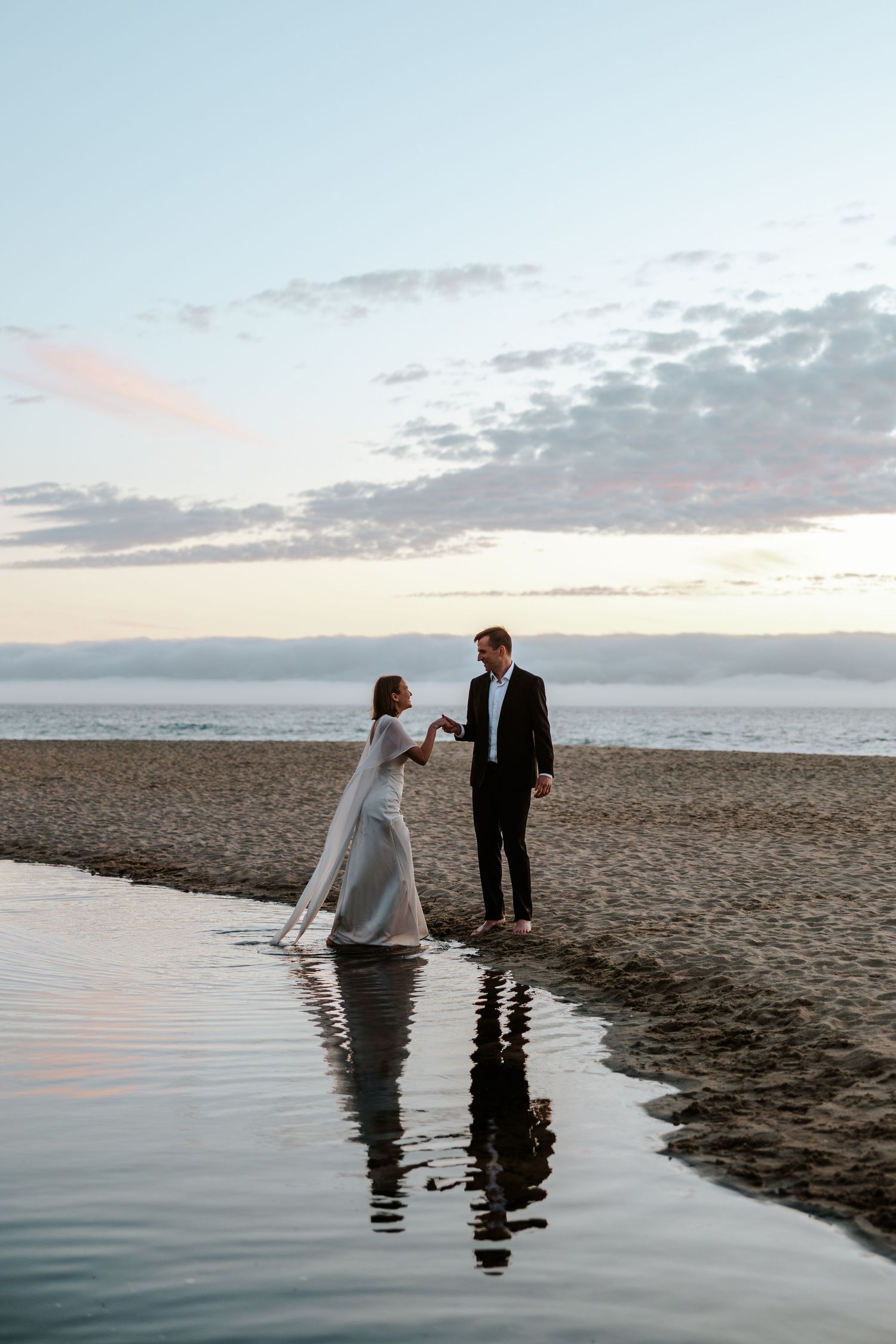 groom helping bride cross pond as the sun sets behind them at the beach and their reflection shows on a pond