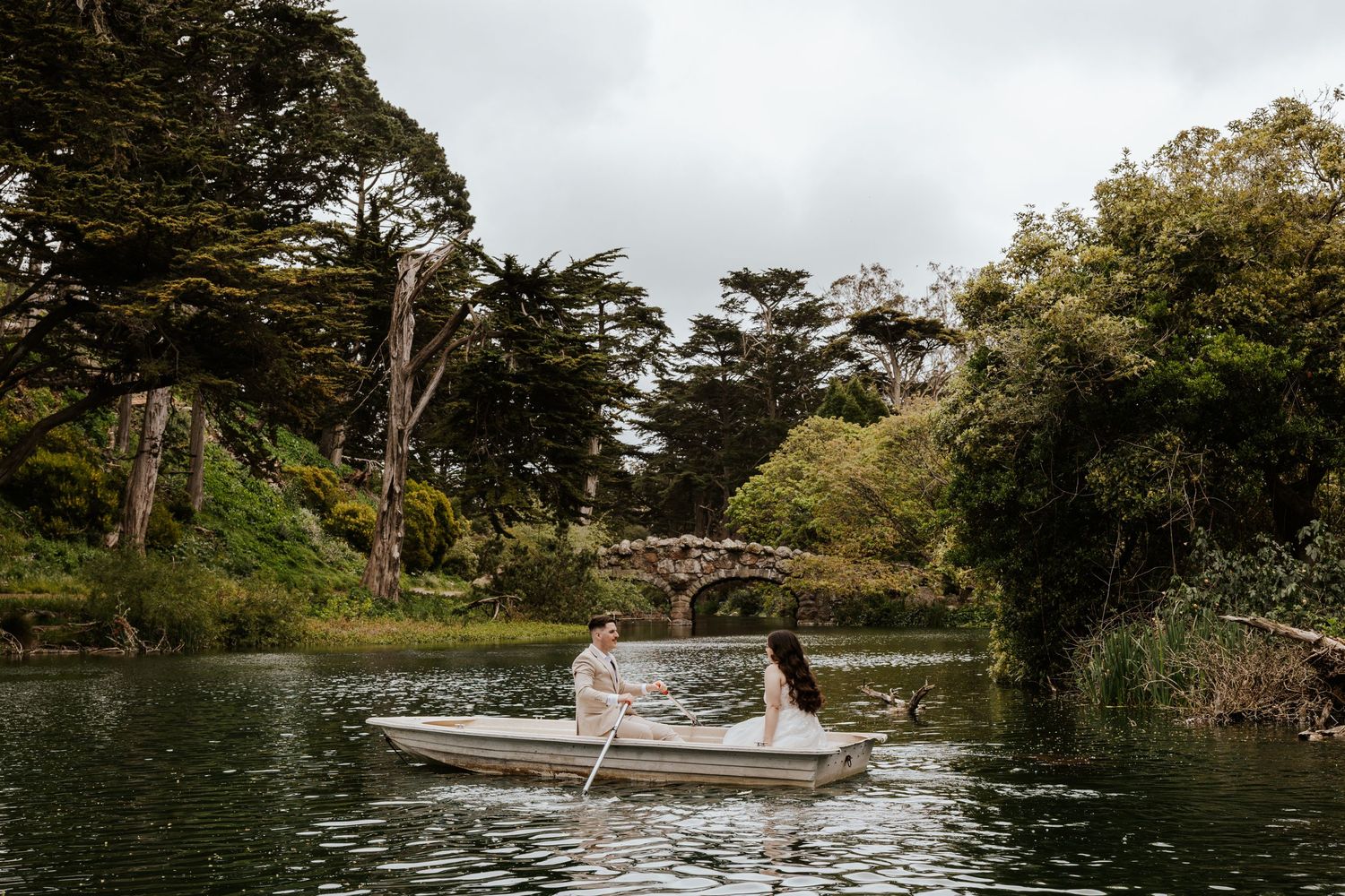 bride and groom in a row boat at stow lake in golden gate park in san francisco during their elopement day with a stone bridge behind them