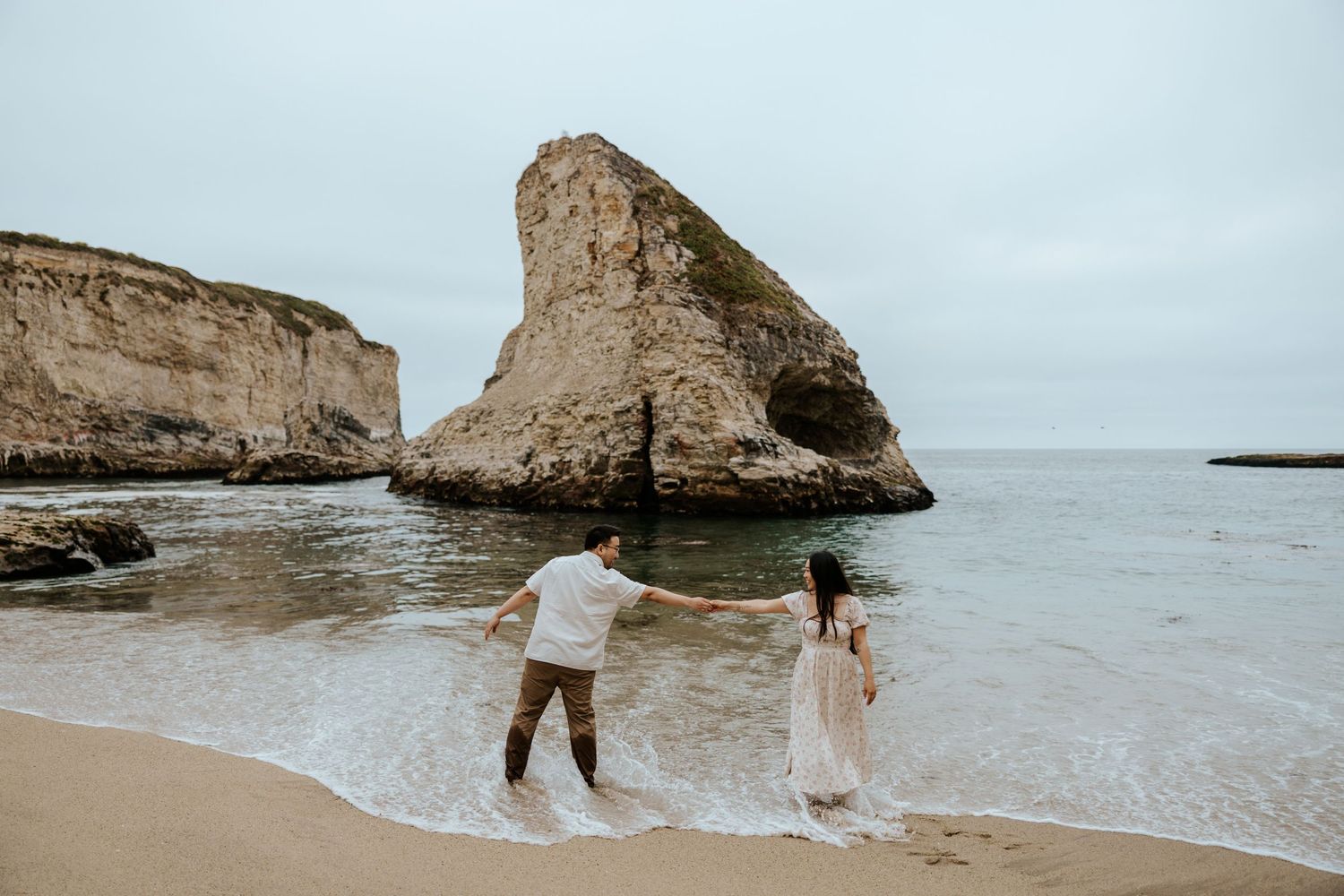 couple playfully running around the beach at shark fin cove in santa cruz during their engagement photoshoot
