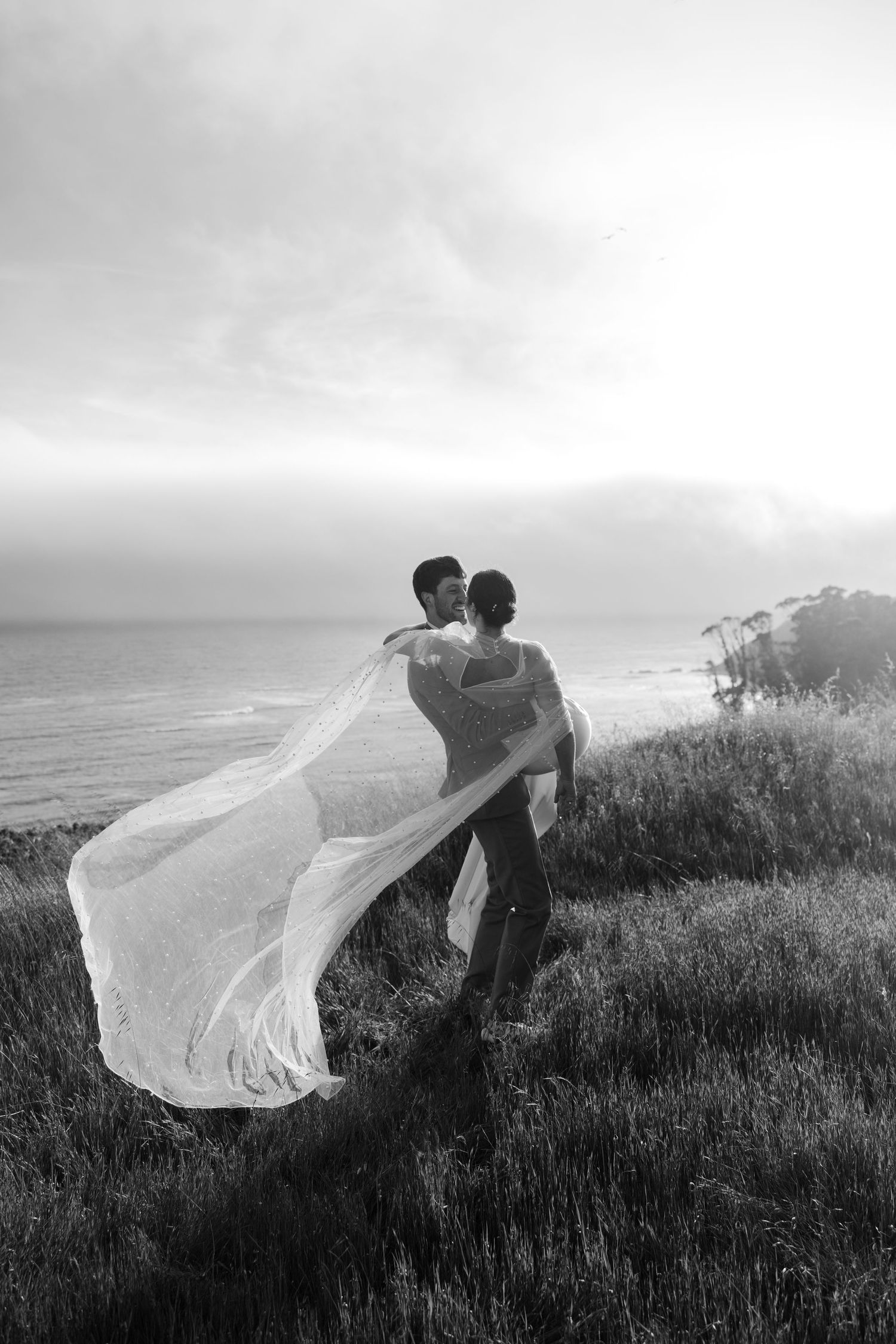 Black and white photo of groom holding his bride on a cliffside while her wedding cape flows in the breeze near San Francisco, California
