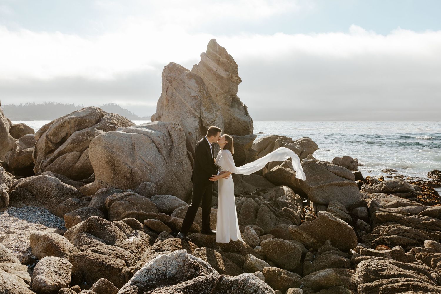 couple kissing as bride's cape flows in the wind while they're standing on giant rocks in carmel, CA