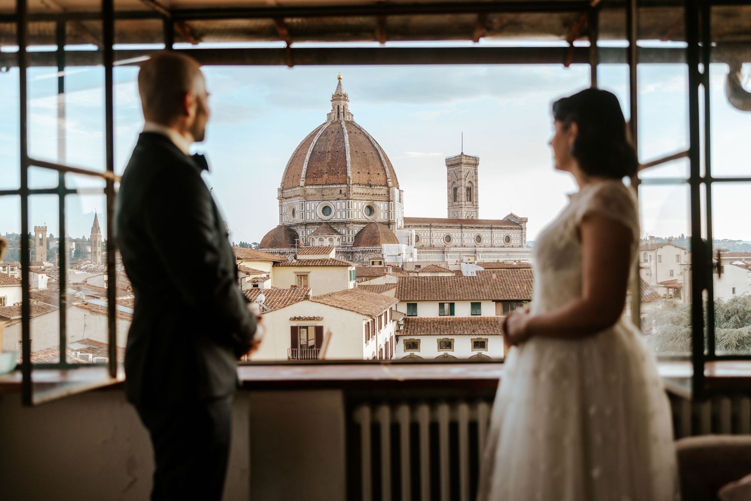 candid couple looking out airbnb florence window towards Duomo on their all day adventure elopement in tuscany, italy