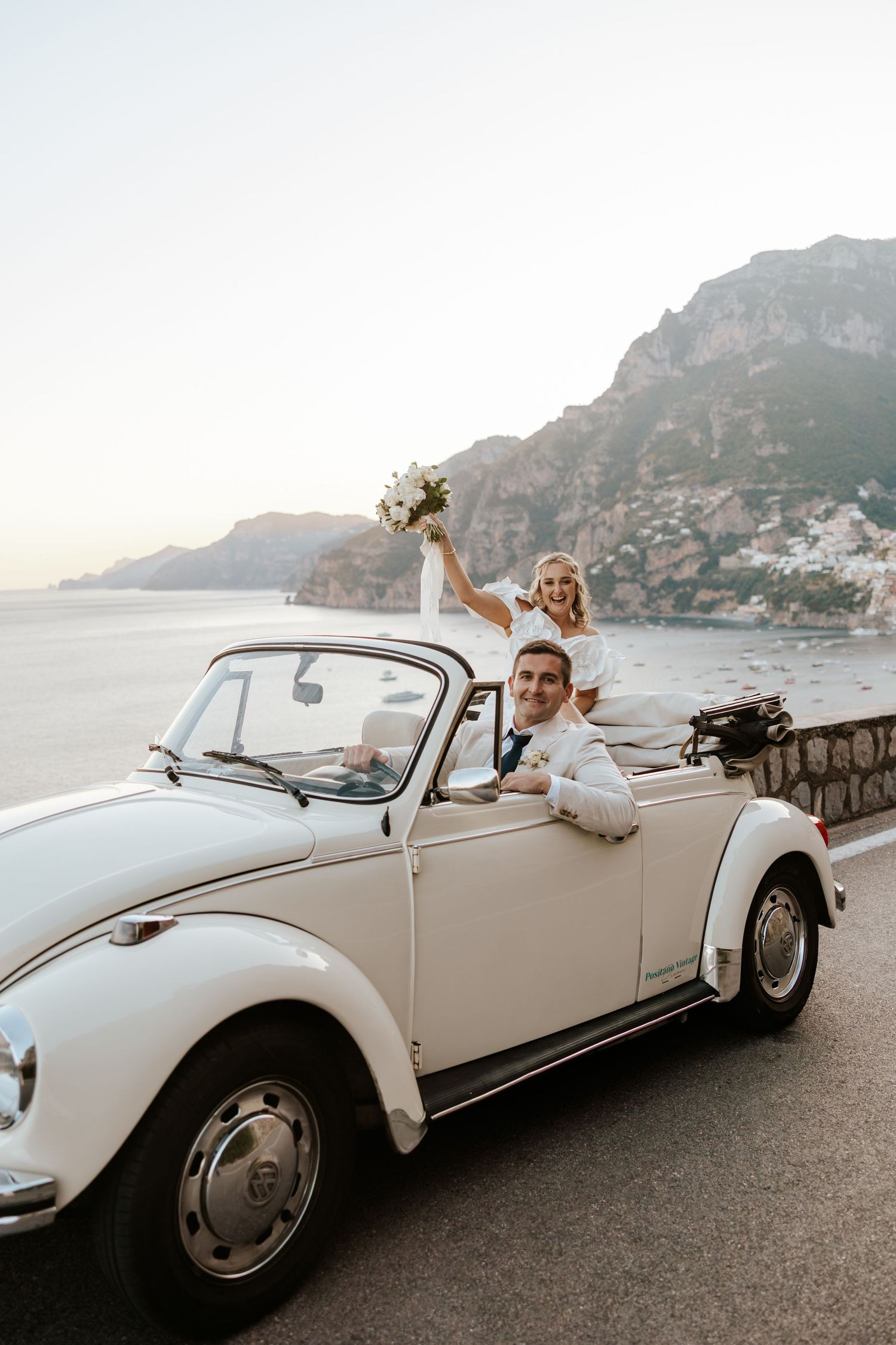 fun bride and groom in a cream vintage car along the cliffs of Amalfi overlooking Positano on their elopement day in Italy