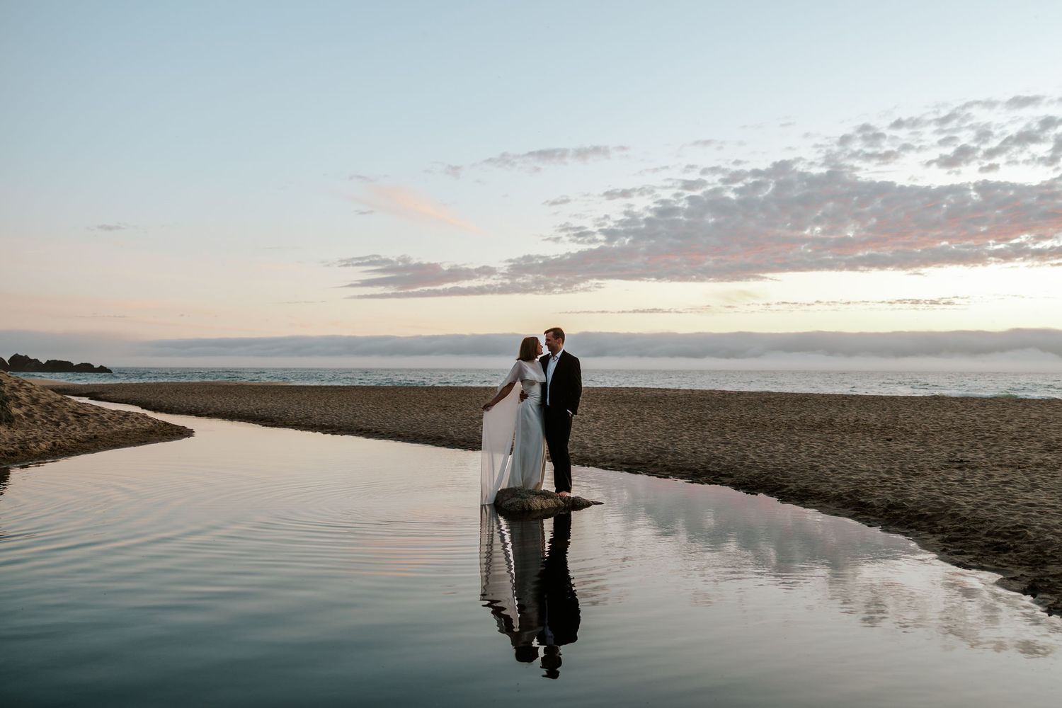 couple embracing as the sun sets behind them at the beach and their reflection shows on a pond