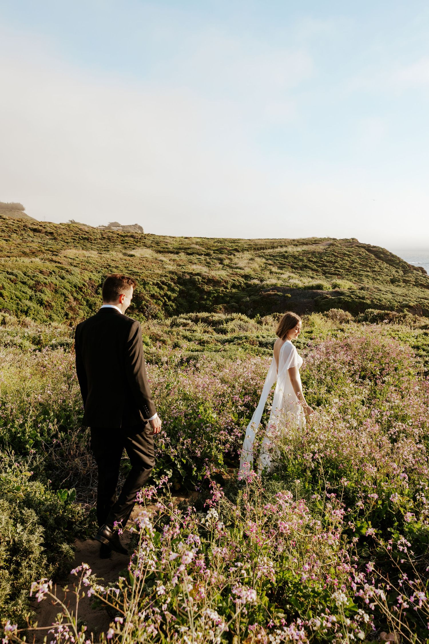 bride and groom walking in a field of flowers on a cliff in big sur