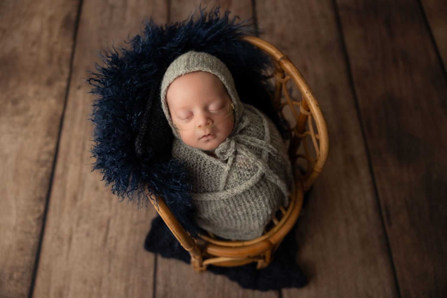 A sleeping baby wrapped in gray knit blanket lies peacefully in wooden basket on rustic wooden floor.