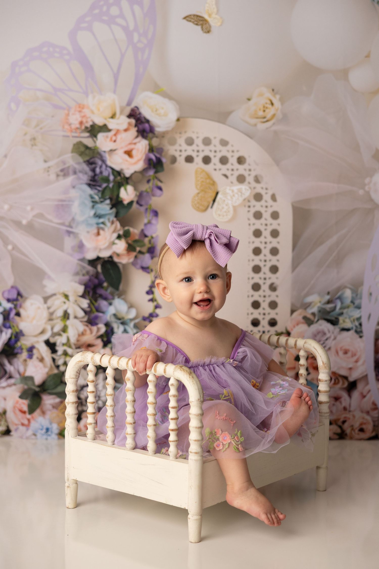 A small bed decorated with floral arrangements in soft purple and white tones creates a dreamy photography setup.