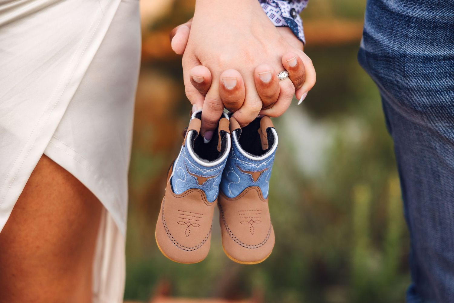 Hands hold small blue and beige baby shoes outdoors in a pregnancy announcement style photo.