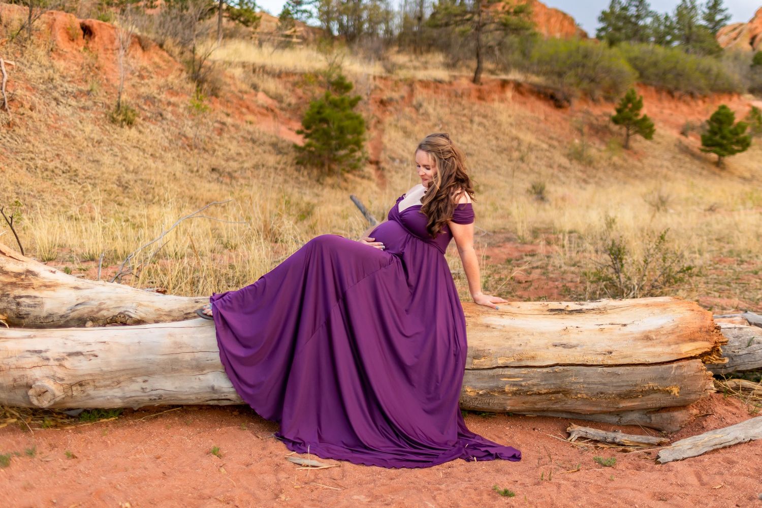 A flowing purple gown drapes elegantly over fallen logs on a rustic hillside during a golden autumn photoshoot.