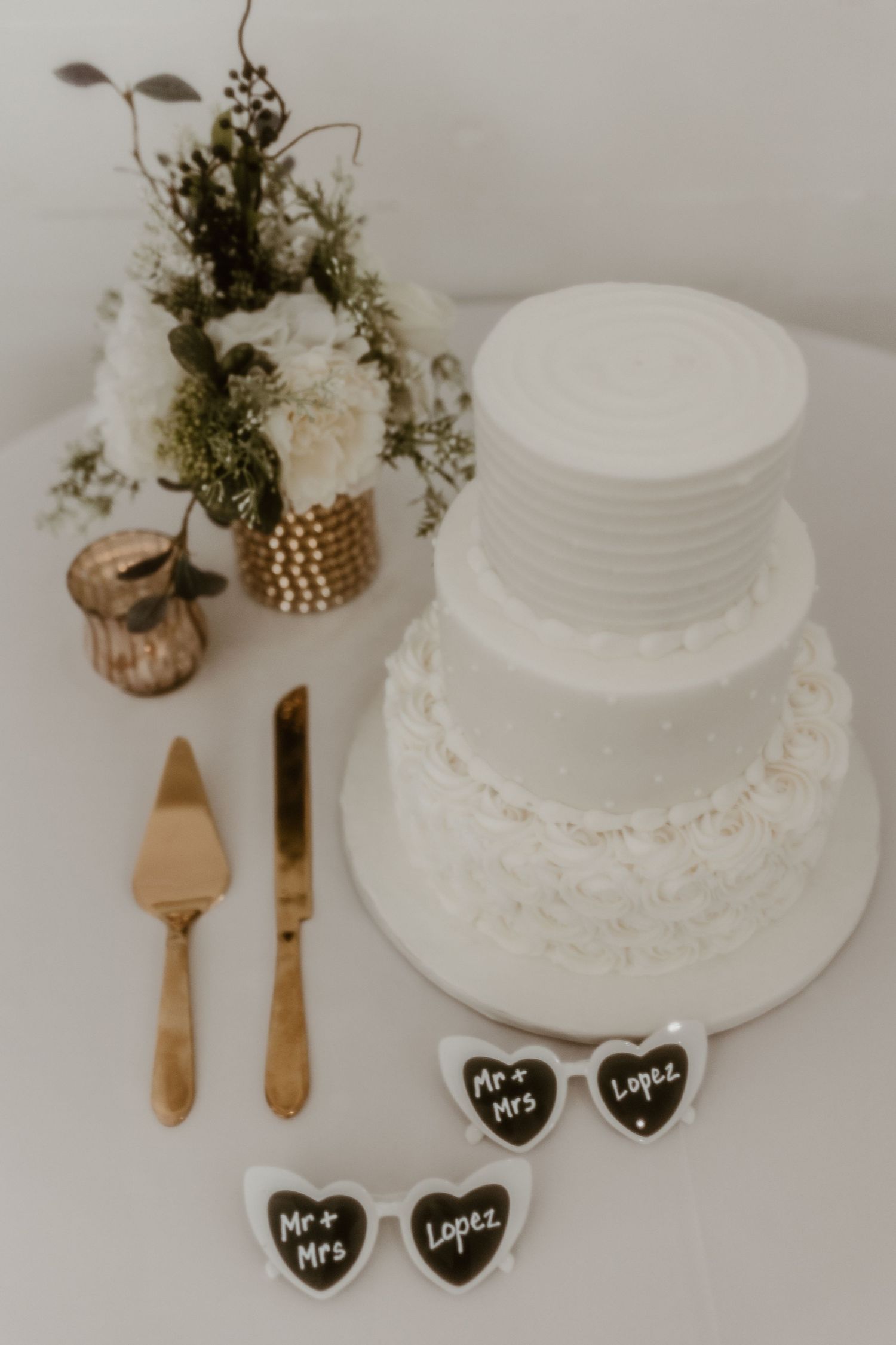 Elegant white wedding cake with gold serving utensils and heart-shaped signs on a table with floral arrangements.