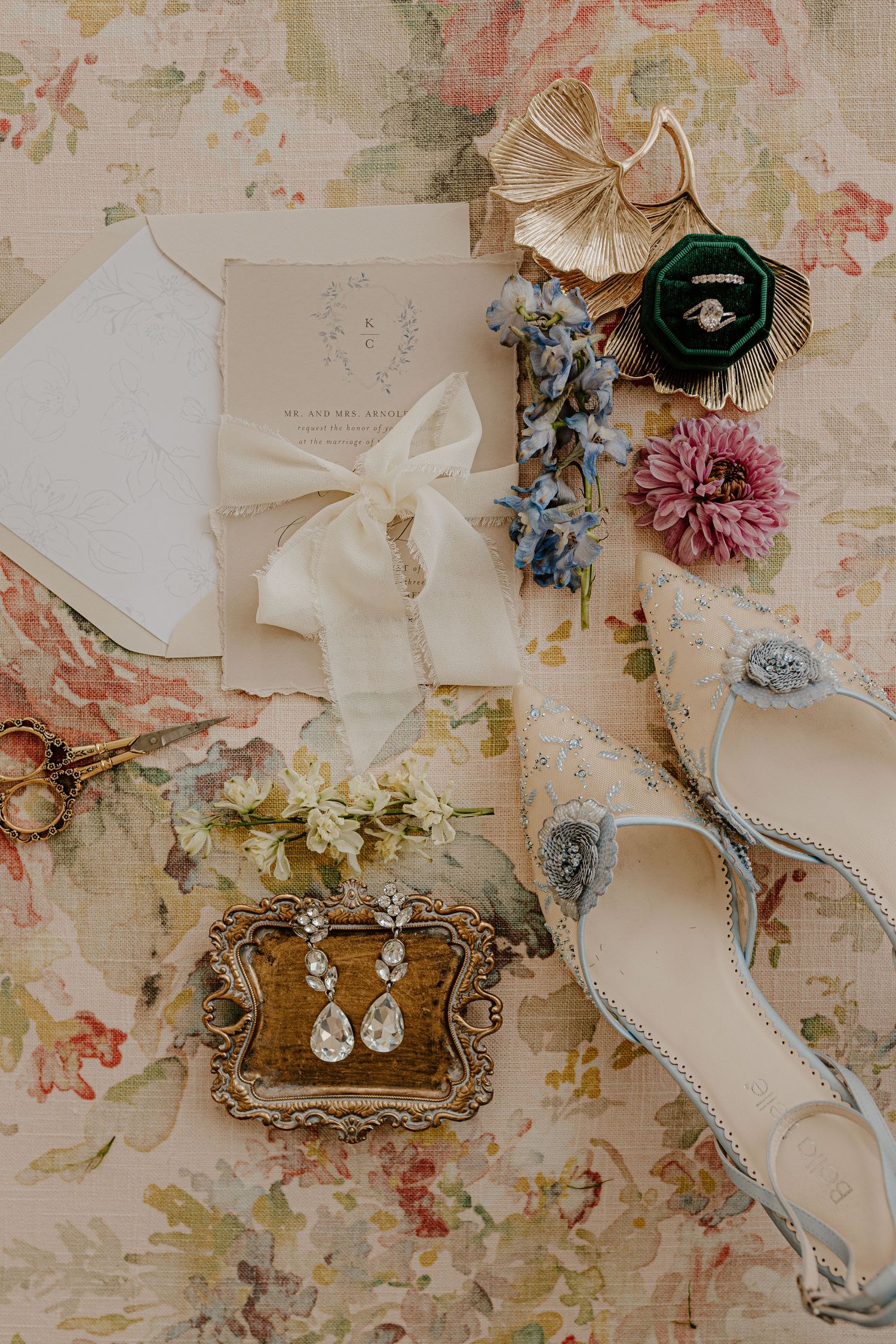 Wedding accessories including vintage earrings, floral accents, and delicate ribbons arranged on floral-print fabric.