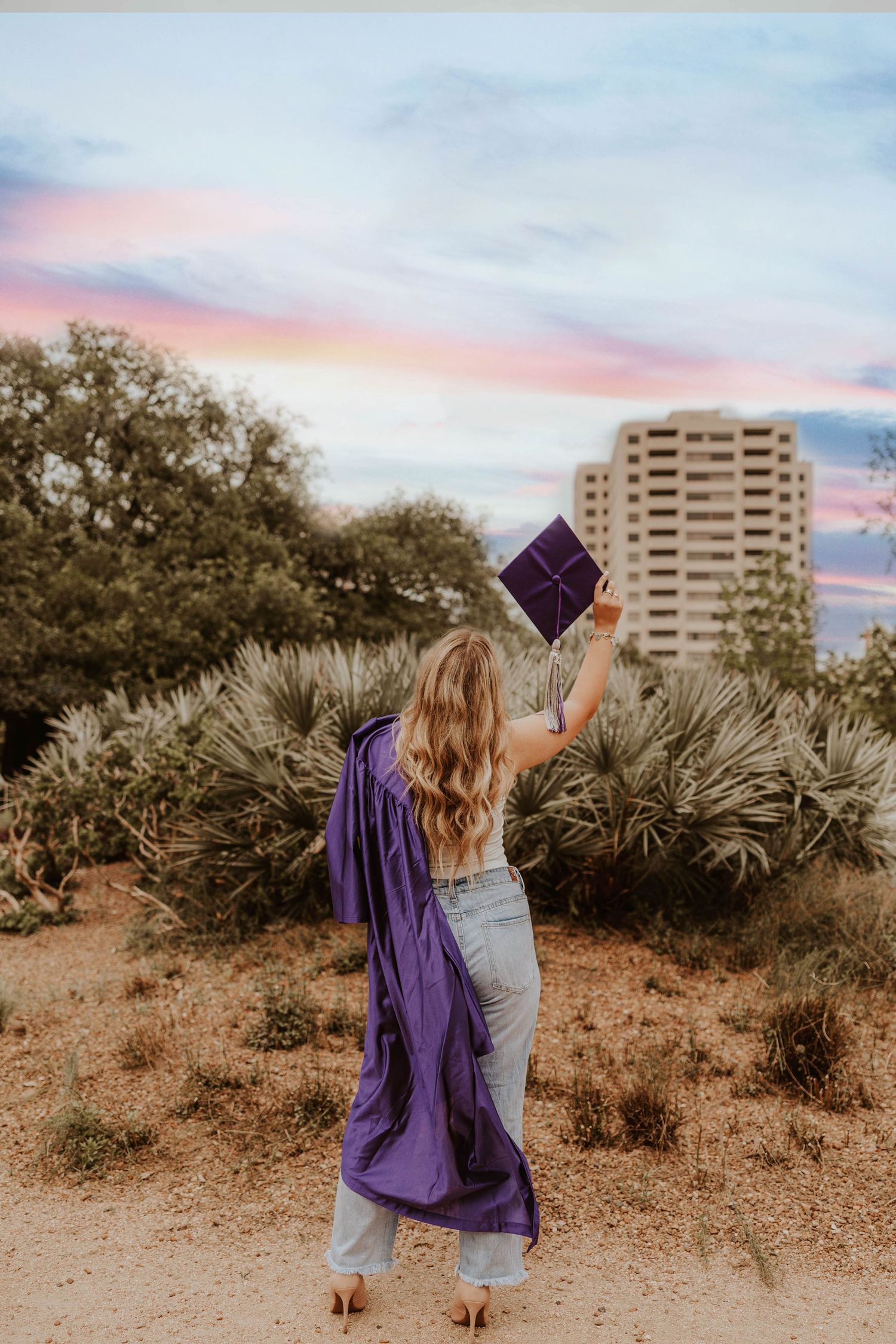 Graduate in purple cap and gown holds up diploma against sunset sky and high-rise building.