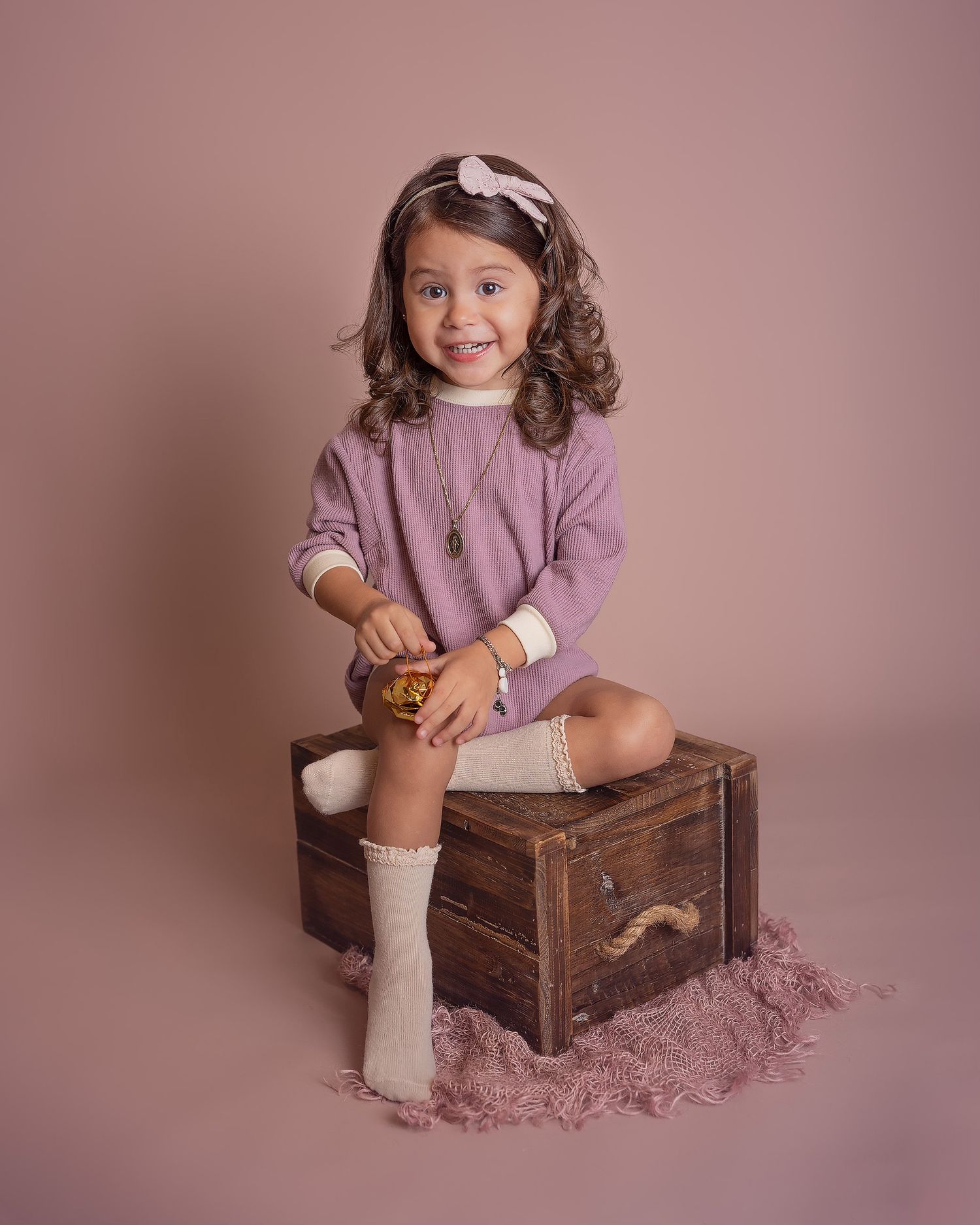A toddler in a purple sweater sits on a rustic wooden crate against a mauve backdrop during a photo session.