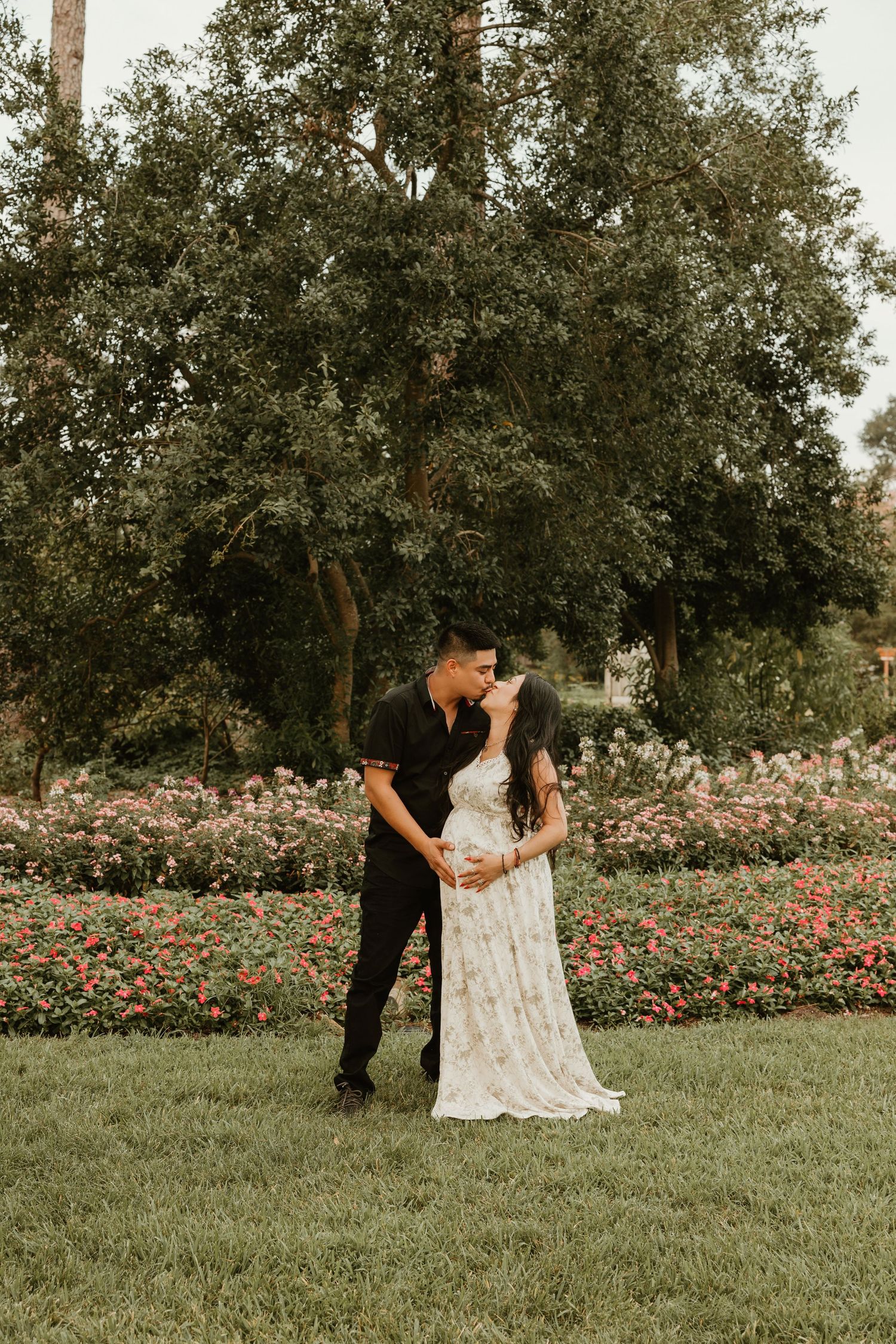 A romantic couple embraces in a garden setting with blooming flowers and tall trees in the background.
