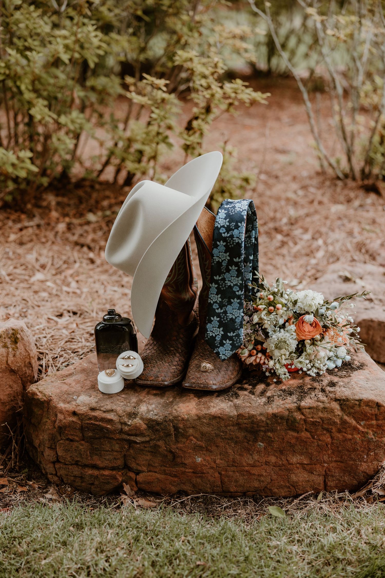 Rustic wedding details with cowboy boots, white hat, and floral bouquet on red rock.
