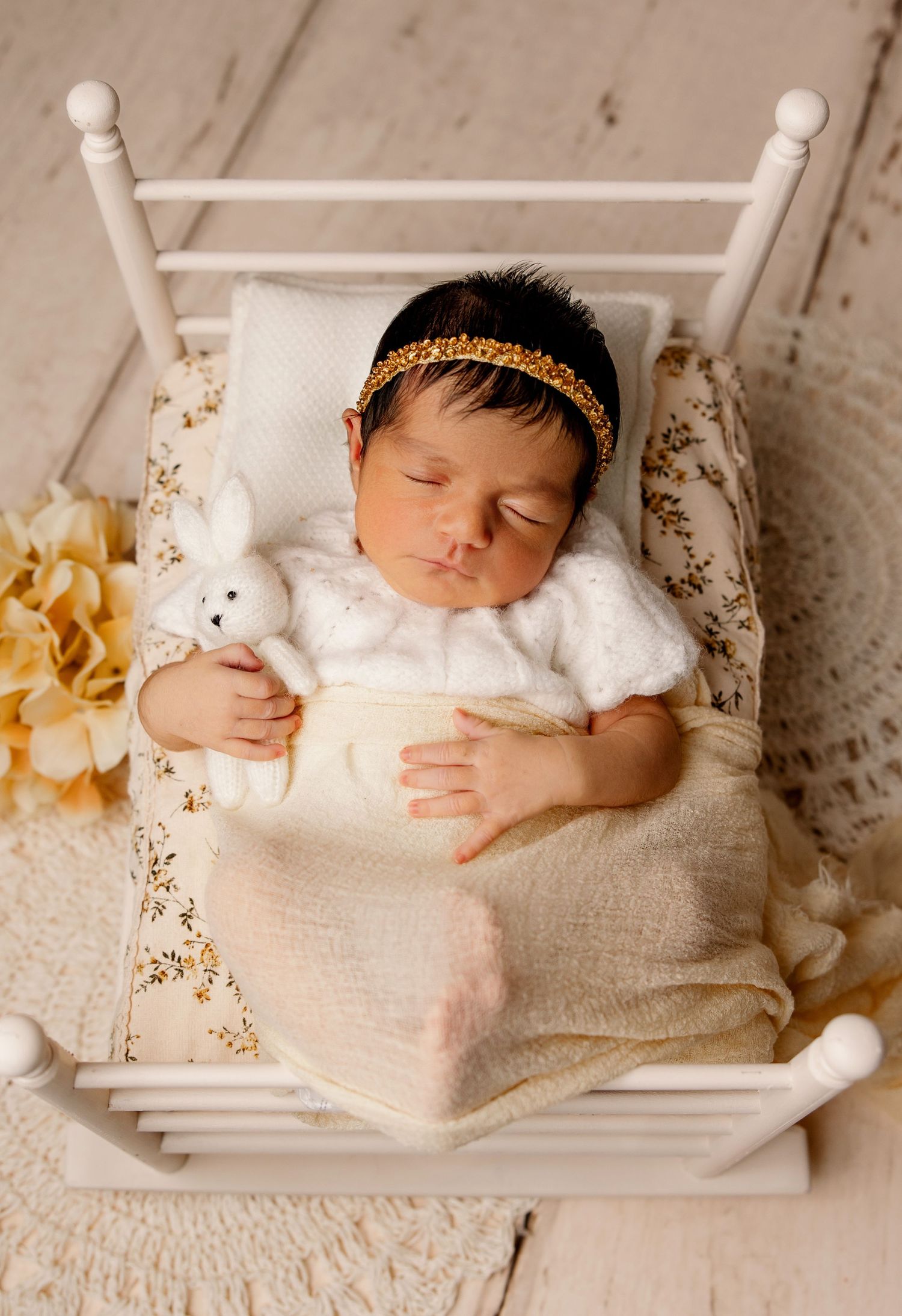 Sleeping baby in white dress with gold headband rests peacefully in decorative white wooden bed with floral accents.