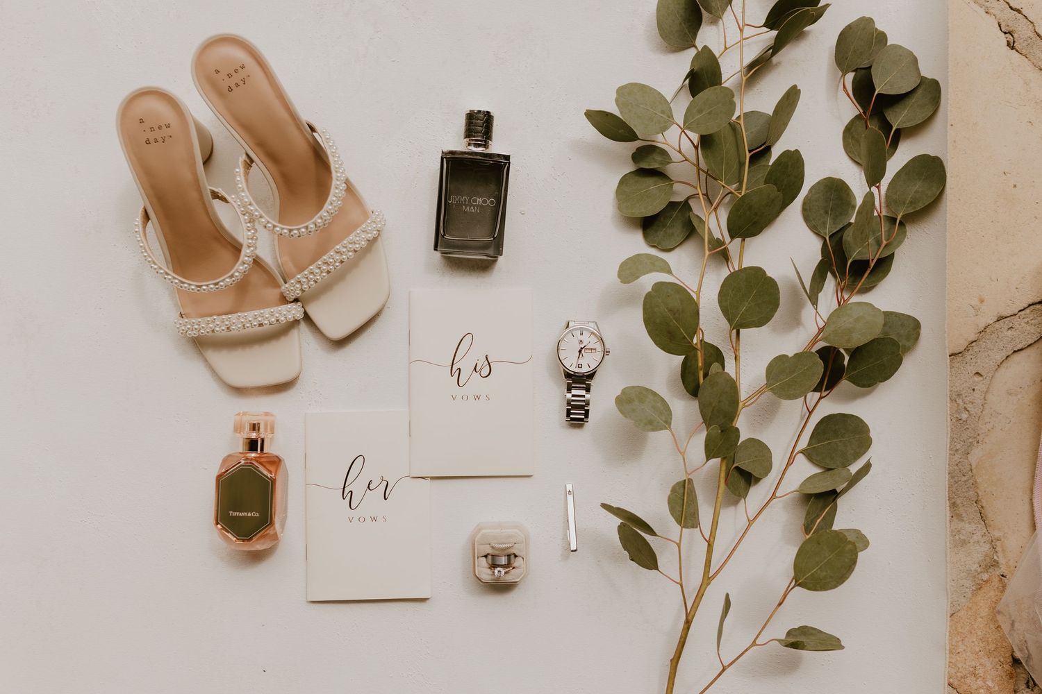 Wedding accessories including shoes, perfume bottles, and vow books arranged with eucalyptus sprigs.