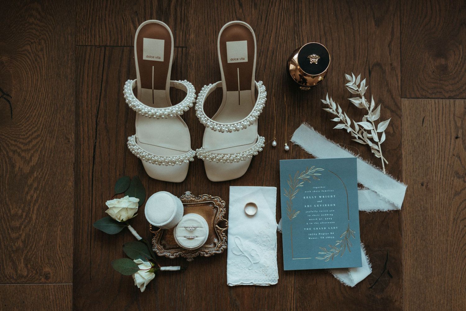 Wedding accessories flatlay with blue invitation, white sandals, and floral details on wood floor.