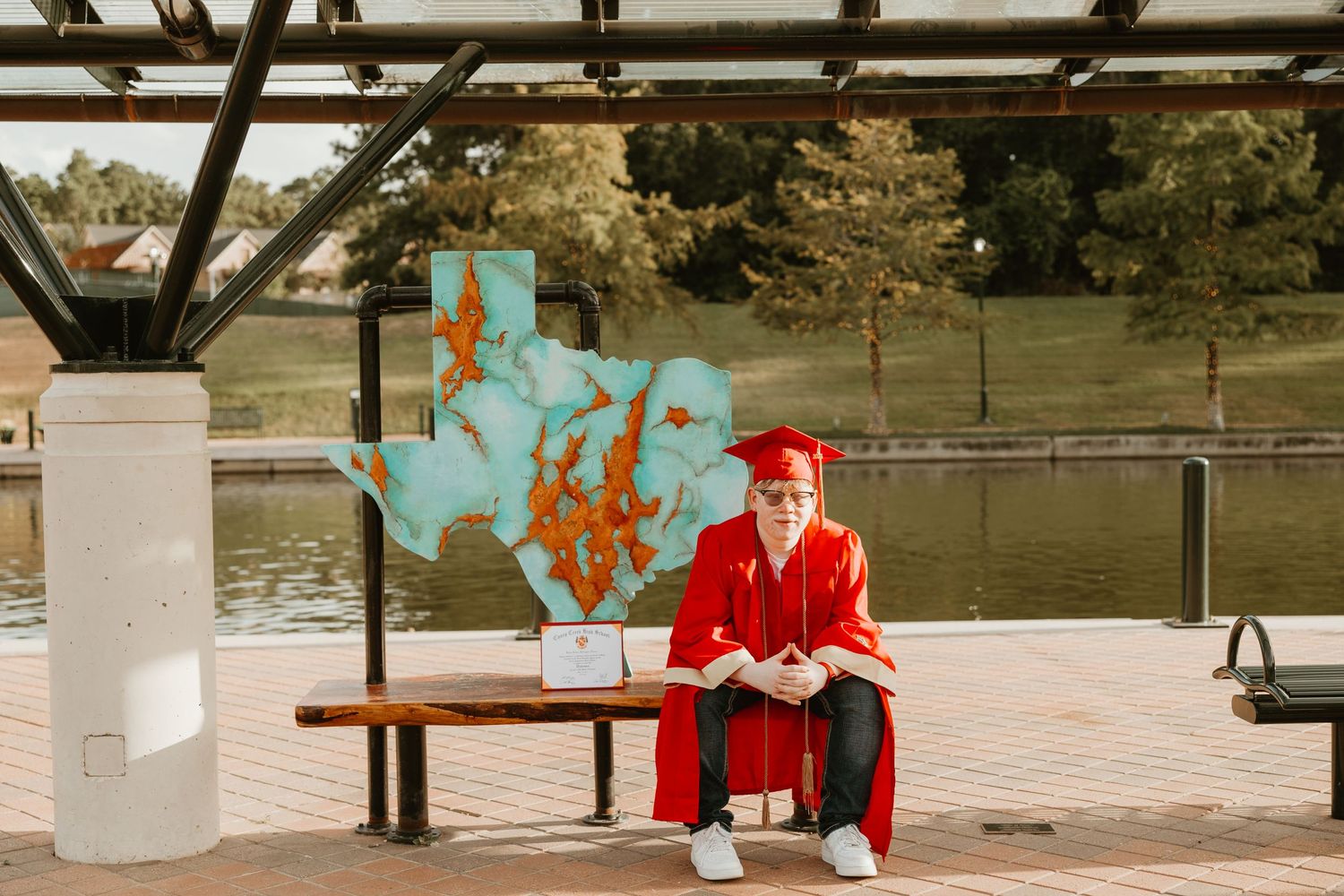 Graduate in red cap and gown sitting on bench near blue wall art outdoors.