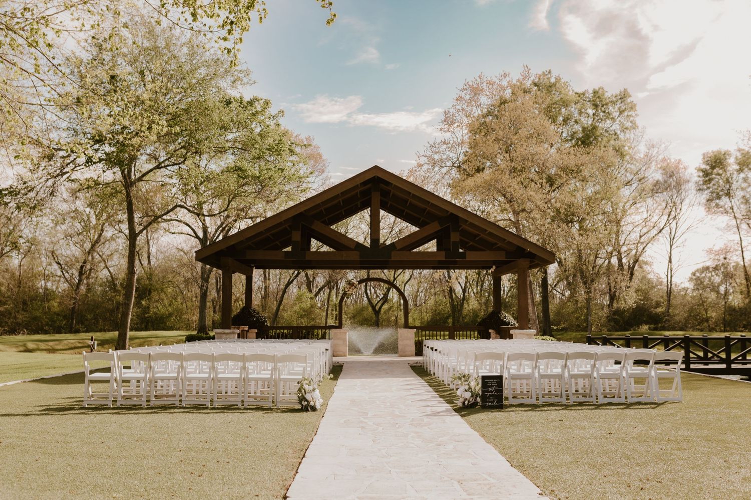 Rustic outdoor wedding ceremony setup with wooden pavilion and white chairs along stone pathway.