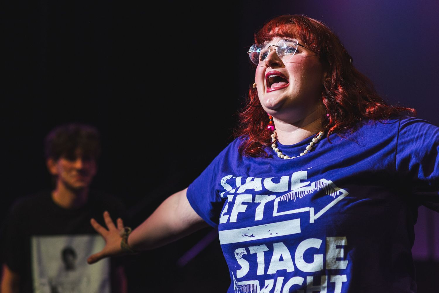 A photo from a production of Be More Chill. There is an actress featured in the front right of the photo with an actor on the left behind.